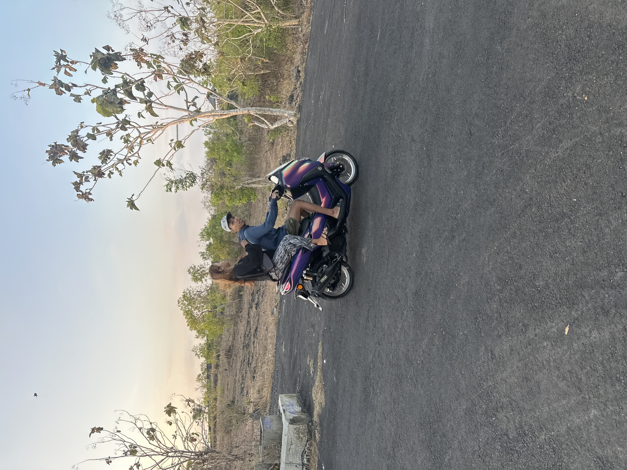 In Kuta Selatan, Indonesia, on a paved road with dry, sparse vegetation on either side, a man and a woman ride a purple scooter. The man, wearing a blue hoodie and shorts, drives the scooter while looking back at the camera with a slight smile. The woman, in a black long-sleeved shirt and patterned leggings, sits behind him, facing forward. The sky is a pale orange and blue, indicating it's either sunrise or sunset. The scooter has a sleek design with iridescent purple and blue panels. To the left of the scooter, there is a concrete structure with some graffiti. The scene is set in a somewhat arid, undeveloped landscape.