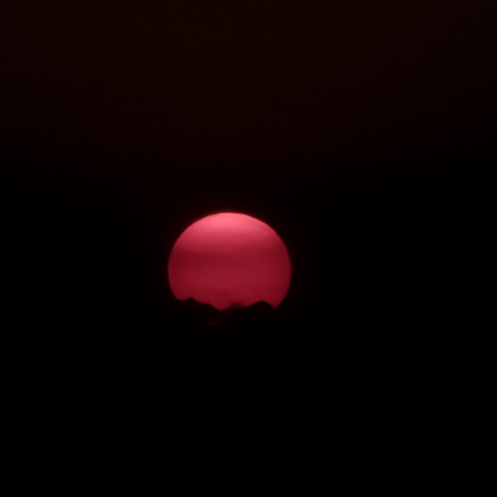 A blood-red sun is partially obscured by a dark, silhouetted horizon of irregular shapes, suggesting mountains or possibly a treeline. The sun is the dominant subject, emitting a soft glow against a pitch-black background. There are no people, structures, or signs of urban life visible. The image appears to be a photograph of a sunset or sunrise, with the dark environment creating a stark contrast with the vibrant color of the sun. The scene is devoid of any context indicating its location in Novi Ligure, Italy, or any other specific place.