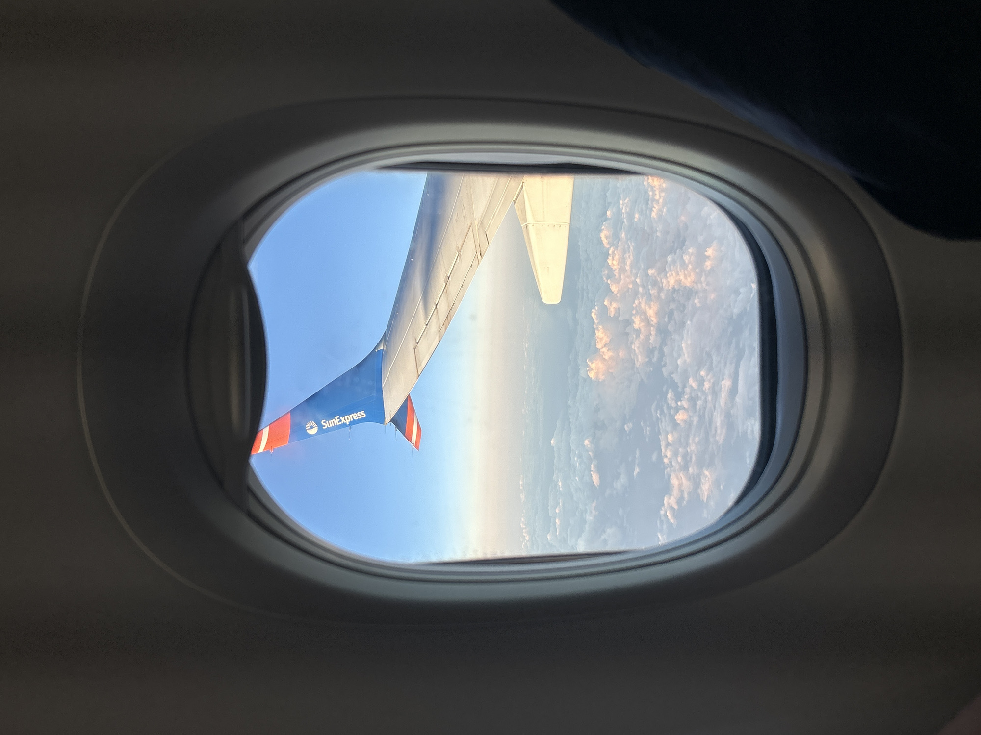 The image, taken from an airplane window, shows a view of the sky above Ambeli, Greece. The airplane's wing and tail fin are visible, with the tail fin featuring the "SunExpress" logo. Below the wing, a layer of clouds stretches across the horizon, tinged with the pink and orange hues of sunrise or sunset. The sky is a clear blue, and the airplane appears to be in flight. The interior of the airplane's cabin is also visible, with the dark interior of the cabin frame around the window. The overall scene conveys a sense of travel and aerial perspective.