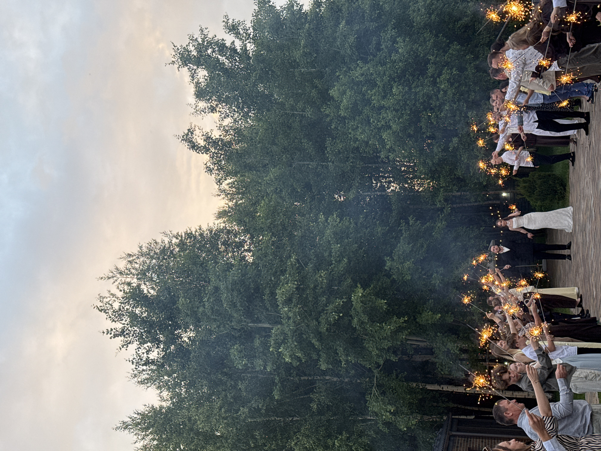 An outdoor celebratory event is depicted at twilight in Perm, Russia. A central couple, a groom in a dark suit and a bride in a white floor-length gown, walks hand-in-hand along a dark paved pathway. They proceed between two lines of approximately 20-30 guests, who hold ignited sparklers aloft, forming a luminous archway. The sparklers emit bright golden-orange sparks and visible smoke. Guests are dressed in a mix of semi-formal and casual attire, including shirts and dresses, and are focused on the central couple. The background is dominated by a dense forest of tall, leafy deciduous trees. A brick building is partially visible on the far left. The sky above presents a gradient of light grey to soft orange, indicative of late evening.