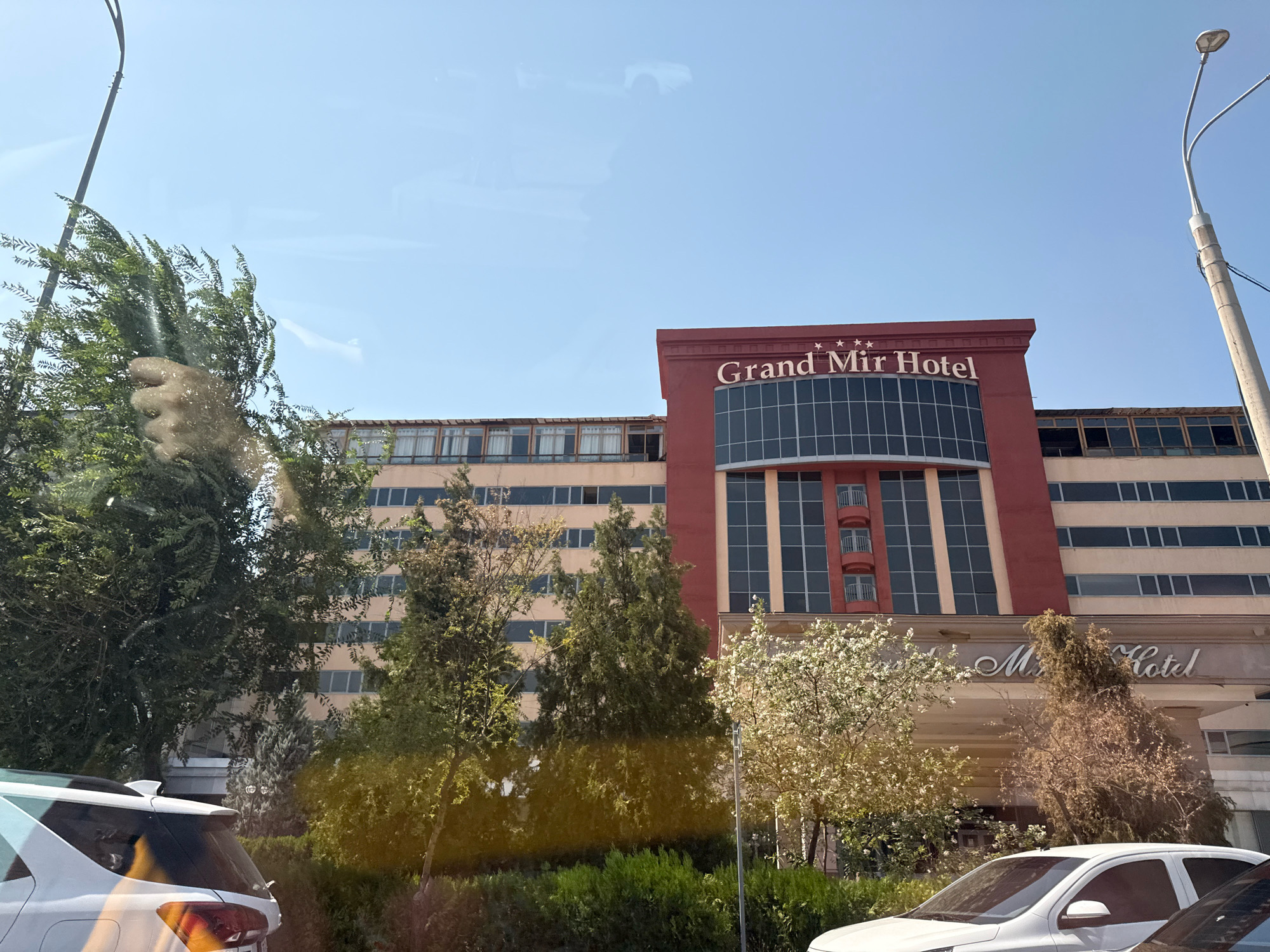 The image captures a view of the Grand Mir Hotel in Tashkent, Uzbekistan, under a clear blue sky. The hotel building is prominently featured, with its distinctive red upper section adorned with the hotel's name and three stars. The lower part of the building is cream-colored, with rows of windows. In front of the hotel, a variety of trees and bushes are visible, some of which are in bloom with white flowers. A white car is parked in the foreground, on the left side of the image. A street light is also visible on the right. The scene appears to be from a street-level perspective, possibly from a vehicle, as indicated by reflections on the windows.