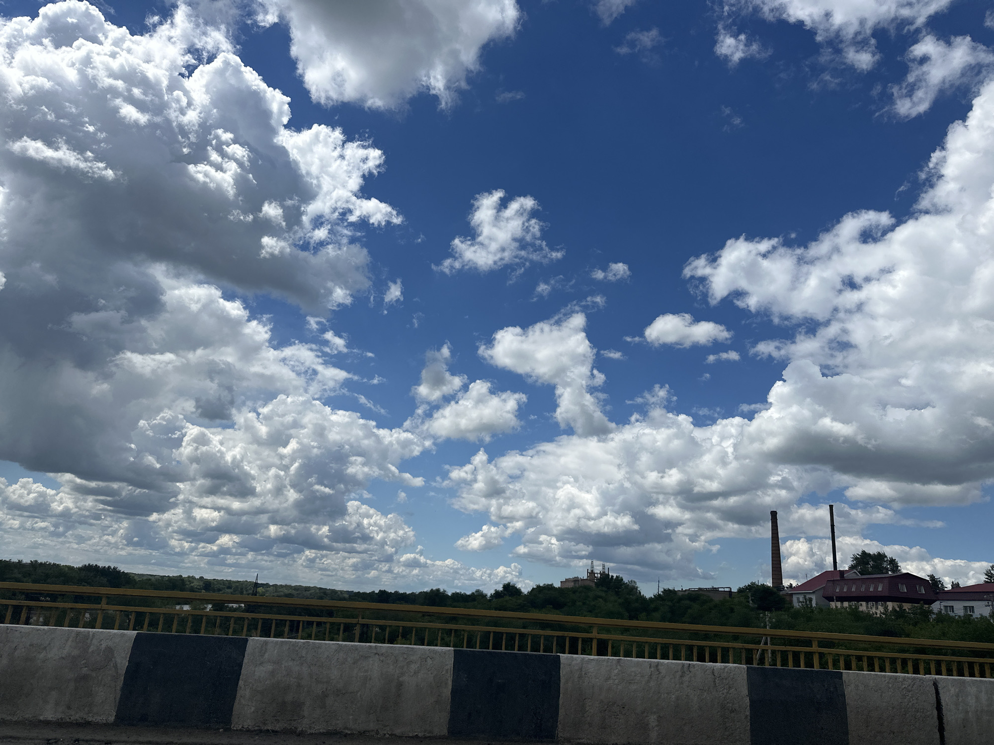 The image depicts a view from a bridge in Alekseyevka, Russia, on a partly cloudy day. In the foreground, a concrete barrier with black and white markings runs horizontally across the bottom of the frame. Beyond the barrier, a yellow railing is visible. The middle ground features a dense line of green trees, and beyond that, several buildings, including what appears to be an industrial complex with two tall, brick smokestacks. The sky is a vibrant blue with numerous large, white cumulus clouds scattered throughout. The overall impression is of a partly industrial, partly natural landscape under a dynamic sky. There are no people or animals visible in the image.