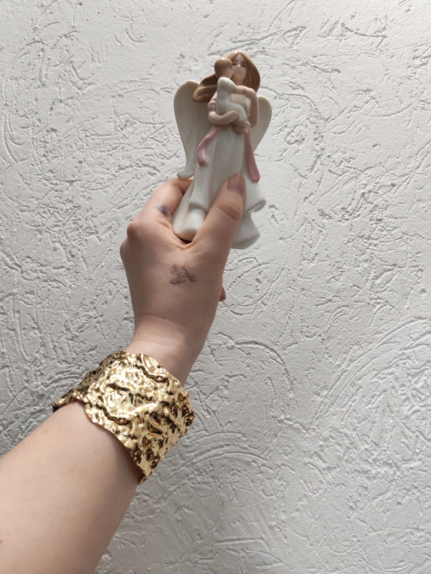 A fair-skinned person is holding a ceramic angel figurine in their hand. The angel is white with brown hair, and is holding a small child. A wide gold bracelet is on the person's wrist. There is a small black tattoo on the back of the person's hand. The background is a white textured wall. The location is not discernible in the image.