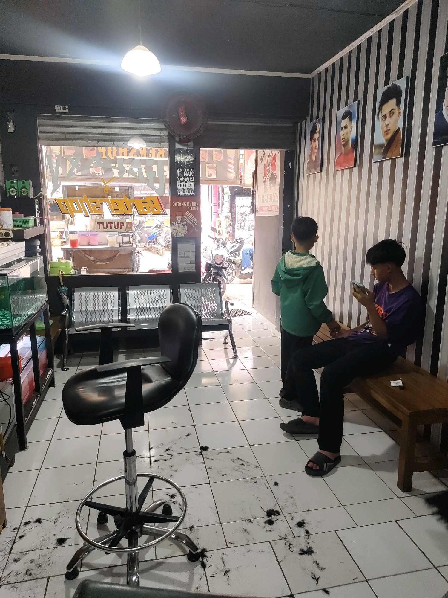 This image depicts the interior of a barbershop in Sukanagara, Indonesia. The scene shows a haircutting station with a black, adjustable barber chair in the foreground, surrounded by fallen hair on the tiled floor. To the right, a wooden bench is occupied by two young males. One, wearing a purple t-shirt and black pants, is seated and looking at his mobile phone. Beside him, another young male in a green hooded jacket stands, with his hand touching the seated individual's arm, suggesting interaction. The background reveals a glass storefront with signage that appears to be in Indonesian, indicating the shop's name and services, including "GANTENG" (handsome). Several framed portraits of young men with stylish haircuts adorn the striped wallpaper on the right wall. Beyond the storefront, the street scene is visible, with motorcycles parked and people passing by, suggesting a bustling urban environment. A ceiling lamp with a white shade illuminates the space. The overall impression is a functional and contemporary barbershop, likely catering to young men and adolescents.