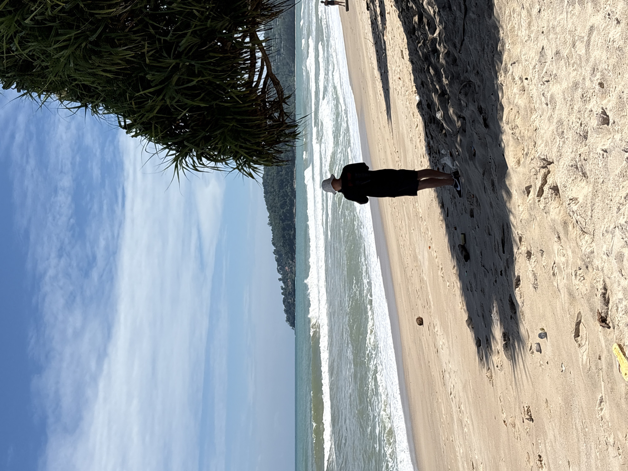 A person wearing a black shirt, black shorts, and a white bucket hat stands on a sandy beach in Choeng Thale, Thailand, facing the ocean. The person appears to be looking at a phone or similar device. The ocean has moderate waves and a distant tree-covered headland. A large, spiky green tree overhangs the right side of the frame, casting shadows on the sand. Other small objects and debris are scattered on the beach, including what appears to be a small rock or coconut, plastic debris, and a yellow object resembling a knife or tool. Another person in dark swimwear is visible further down the beach. The sky is blue with scattered white clouds.