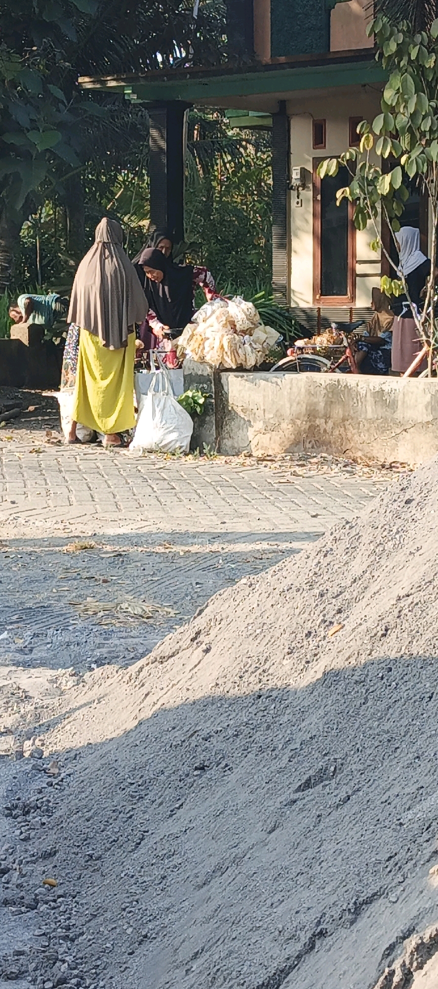 In Kasiyan, Indonesia, a group of individuals are gathered near a building with a veranda, possibly a home or shop. The scene appears to be outdoors, with vegetation visible in the background and a pile of sand or gravel in the foreground.
Three women are the primary subjects in the image. One woman, on the left, is wearing a light brown hijab and a bright yellow skirt. Another woman, in the center, is dressed in a black hijab and a patterned top. The third woman, to the right of the center, is also wearing a light brown hijab and a dark skirt. The women are standing around a collection of items, including what appears to be a large mound of white material, possibly fabric or bundled goods, and a white shopping bag. A bicycle is partially visible behind the mound of items.
The ground in front of the building is paved with rectangular stones. The environment suggests a warm climate, with sunlight casting shadows. The overall impression is of a community gathering or a marketplace scene, with people engaged in commerce or social interaction.