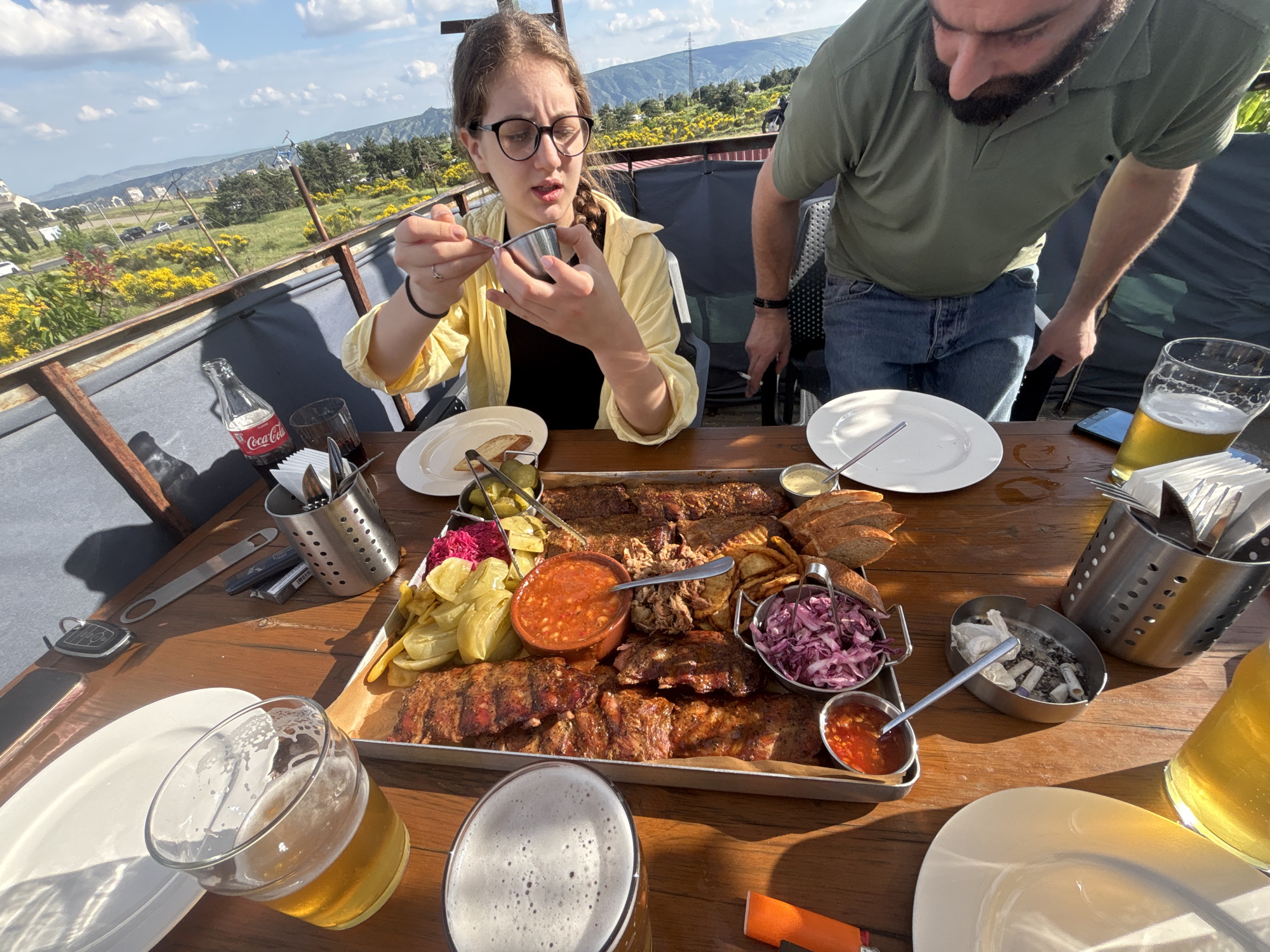 The image depicts a group of people enjoying a meal at an outdoor restaurant overlooking a scenic landscape. In the foreground, a large metal platter is filled with various food items, including ribs, pulled pork, coleslaw, french fries, and sauces. Several plates, glasses of beer, and cutlery are arranged on a wooden table.
A young woman with glasses and braided hair is seated at the table, holding a small metal container in her hands. To her right, a man with a beard is standing, wearing a green polo shirt and jeans. The background reveals a panoramic view of Tbilisi, Georgia, with rolling hills, urban structures, and a clear sky with scattered clouds. The restaurant appears to be situated on an elevated terrace, offering a picturesque vista of the city and its surroundings. The overall scene suggests a relaxed and enjoyable dining experience.