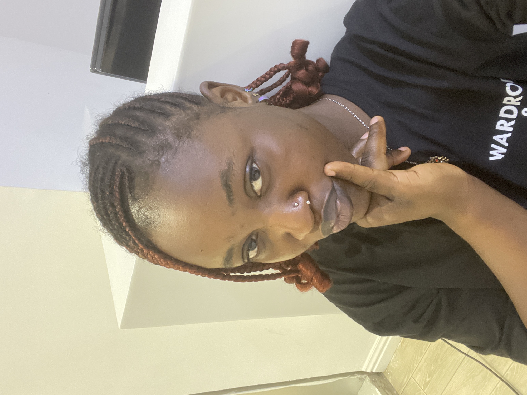 A medium shot captures a young Black woman from the chest up, positioned indoors. She is looking directly at the camera, her right hand resting on her chin, with her index finger extended towards her lips. The woman has braided hair with reddish-brown accents, some braids styled down, and others secured in buns. She is wearing a black t-shirt with white text visible on the lower right portion, partially obscured. She has a nose piercing with two small studs and is wearing a thin silver chain necklace with a pendant. The background appears to be an indoor setting with light-colored walls and a white corner structure. The floor is tiled with a light beige color, and a black cable is visible on the floor. There is a hint of a dark rectangular object on a shelf in the upper right corner of the image. The overall lighting suggests an indoor environment, possibly during the day. The setting is not definitively identifiable as Unguwar Sarki, Nigeria, based solely on the visual information provided.