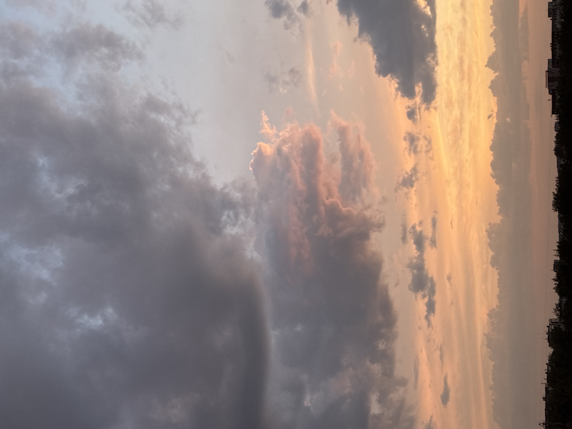 The image depicts a wide shot of a twilight sky over an urban landscape. The sky is dominated by dramatic clouds in varying shades of gray, pink, and orange, suggesting the aftermath of a sunset or an approaching storm. Towards the bottom of the frame, a dark silhouette of trees and buildings lines the horizon, indicating a city or town. There are no people or discernible actions visible in the scene, the focus being on the atmospheric conditions of the sky and the subdued urban edge. The overall impression is one of natural beauty juxtaposed with human development.
