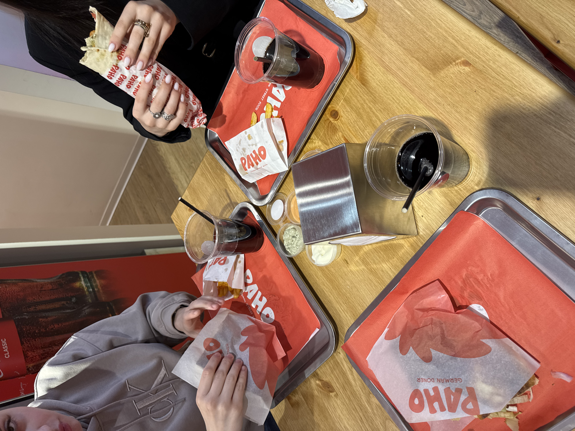This is an eye-level, medium shot of a table in what appears to be a restaurant or fast-food establishment, likely in Yerevan, Armenia. Two individuals are partially visible, each holding a "PAHO German Doner" sandwich.
One person, wearing a black jacket, is seen in the upper left corner, holding a sandwich wrapped in printed paper. The other person, wearing a gray hoodie with the letters "ME" embroidered, is near the bottom left corner.
On the wooden table, there are three metal trays lined with red paper, each containing a wrapped sandwich with "PAHO German Doner" branding. Two plastic cups with dark liquid and straws are also on the table, along with a small bag of fries and several small condiment containers. A metal napkin holder is positioned between two of the trays.
A Coca-Cola advertisement is partly visible along the left edge of the image, further suggesting a restaurant environment. The overall setting suggests a casual dining experience, possibly in Yerevan, Armenia.