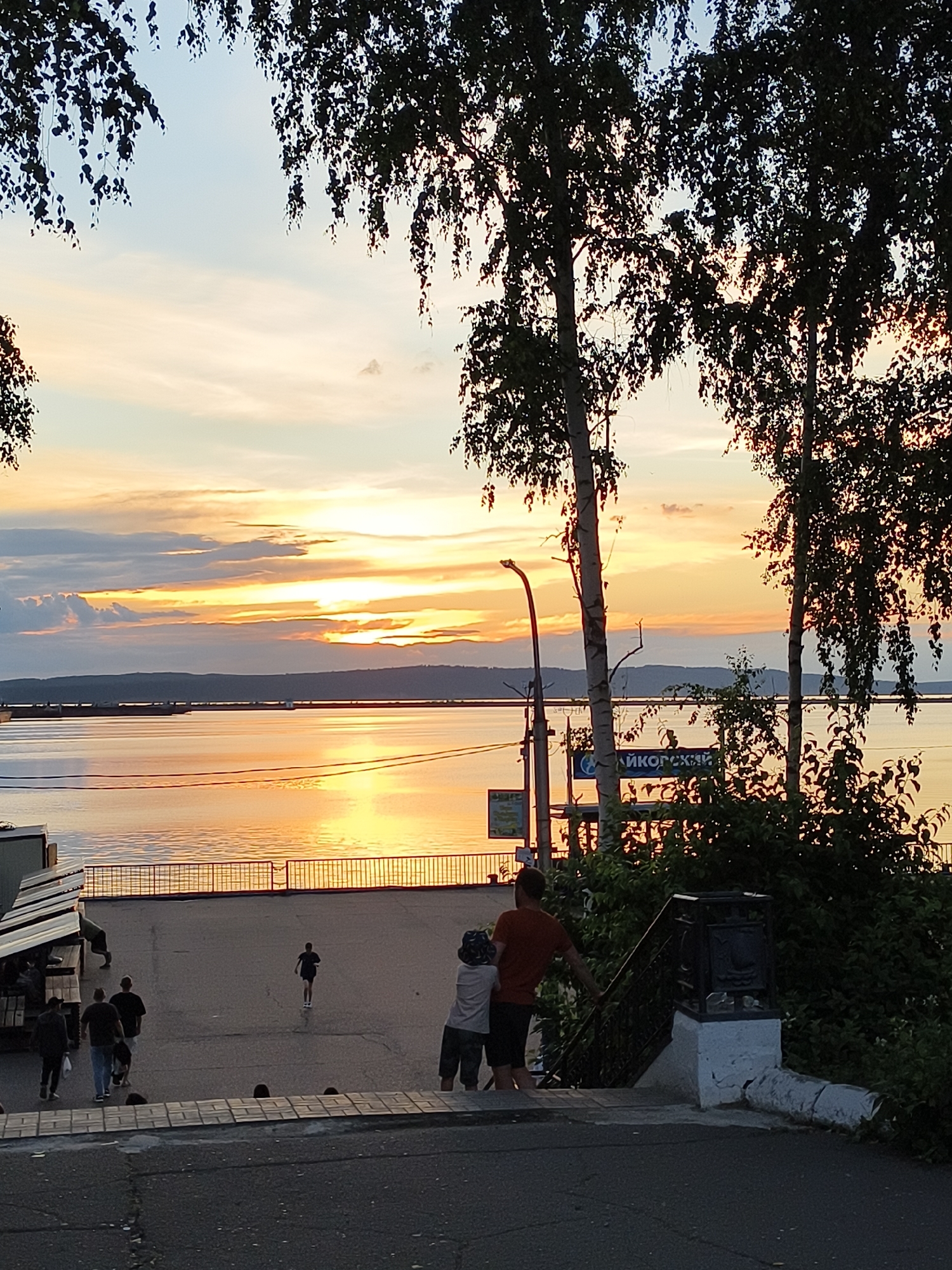 This media file captures a scenic sunset over a body of water. The sky is ablaze with warm hues of orange, yellow, and pink, reflecting beautifully on the calm surface of the water. Silhouetted trees with lush foliage frame the top of the image, their leaves creating delicate patterns against the colorful sky.
In the foreground, a paved area leads down towards the water. Several people are visible on this pathway, some walking, others gathered. A few individuals are walking towards the water, while others are conversing. In the middle ground, a young boy runs across the path. Further in the background, along the waterfront, there are some structures that appear to be market stalls or small shops, with a railing separating the paved area from the water.
A streetlamp stands on the left side of the paved area, and a sign with Cyrillic lettering is visible on a post near the water's edge. The sign appears to read "ЯЙКОРАНСКИЙ" which could be a place name or part of a business name. The overall atmosphere suggests a relaxed evening setting, possibly a promenade or a public space by a river or lake. The warm colors of the sunset evoke a sense of peace and tranquility. The time of day is clearly sunset, and the weather appears to be clear, conducive to outdoor activities.