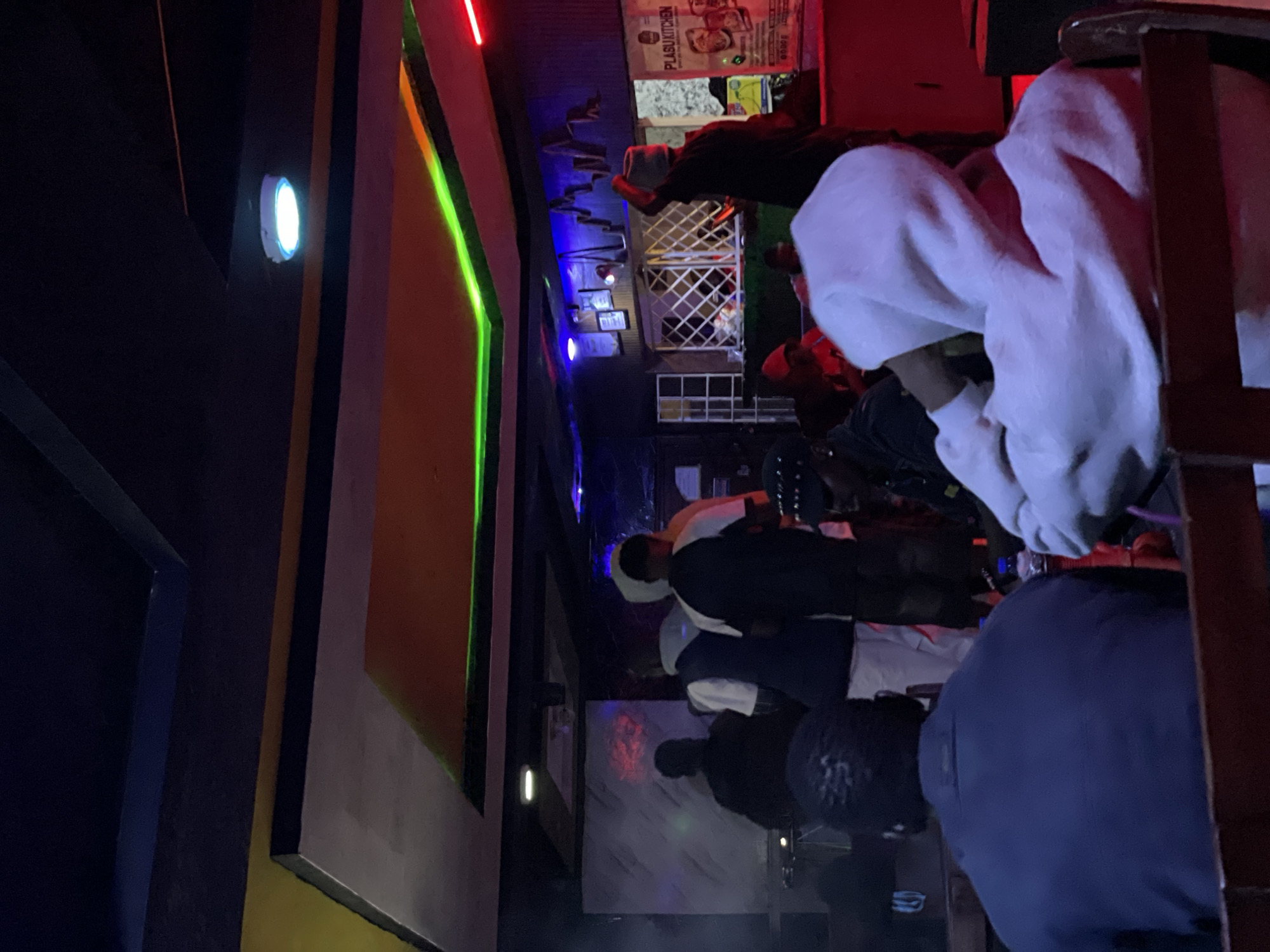 The image captures an indoor scene, likely a nightclub or a bar, with dim, colorful lighting. In the foreground, a person wearing a blue jacket is seated with their back to the camera, facing a red-brown wooden surface. To their right, another individual in a white hoodie is also seated, facing away. The background features several people standing, some appearing to be engaged in conversation or observing an area off-camera. One person is visible in a black shirt and a grey hat, holding their arm up. Further back, a counter area with a patterned grille is present, with framed items displayed on the wall behind it. A poster with text, including "PLASU KITCHEN," is visible on the right side. The ceiling is illuminated with colored LED lights in green and orange hues, and a white circular light fixture is present. The overall atmosphere suggests a social gathering or entertainment event. The specific location is a commercial establishment in Durumi, Nigeria, characterized by its nightlife and entertainment.