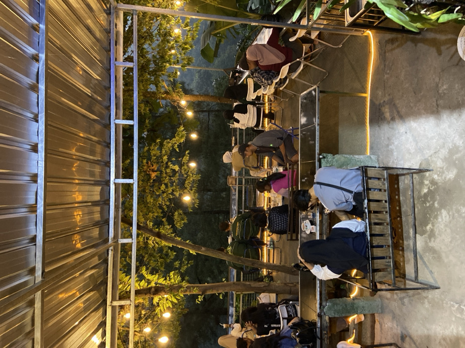 The image depicts a nighttime scene at an outdoor or semi-outdoor establishment in the city of Cembor, Indonesia, likely a cafe or restaurant, illuminated by artificial lighting. A corrugated metal roof spans the top, supported by a metal frame. Beneath the roof, multiple string lights with exposed bulbs are strung between structural elements and through the foliage of several trees, casting a warm glow on their leaves.

Approximately 15-20 individuals are present, seated at various tables and benches. In the foreground, a man in a grey shirt and a woman in a black hijab and white long-sleeved shirt are seated on a wooden bench, facing away from the camera, at a table with a dark surface. Nearby, a yellow LED strip light outlines a section of the concrete floor.

In the middle ground, groups of people are seated at multiple tables. One table shows a young girl in a pink top and another individual. To their left, a person in a green and white long-sleeved shirt sits at a table near the background railing. To the right, individuals in hijabs are seated at tables with white folding chairs. A man in a grey shirt and cap sits facing left.

In the background, beyond a metal railing, dense foliage and trees are visible against the dark night, suggesting a natural, elevated setting. The overall atmosphere appears social and relaxed. Objects on tables include cups, glasses, and small dishes.