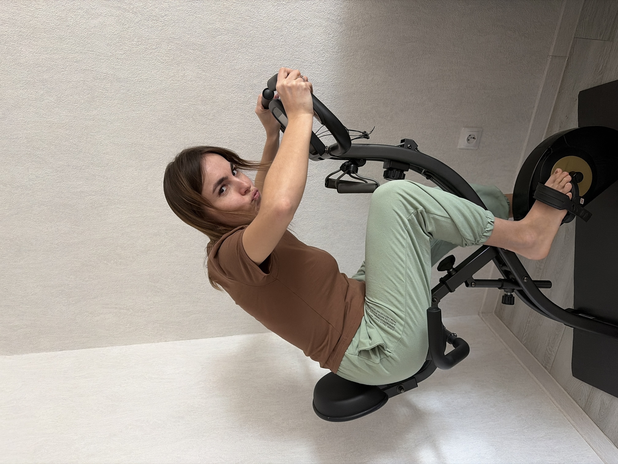 A woman is working out on an exercise bike. She is wearing a brown t-shirt and light green sweatpants. The location appears to be indoors, with light-colored walls. The visible text on the sweatpants includes "THESE TRACK PANST ARE A RECYLED WATER ENVIRONMENTALLY FRIENDLY DYE CRE FROM A RECYLED ANG ORGAN WATER BY." There is an electrical outlet visible on the wall. There is no information given in the image to establish the location as Karyakino, Russia.