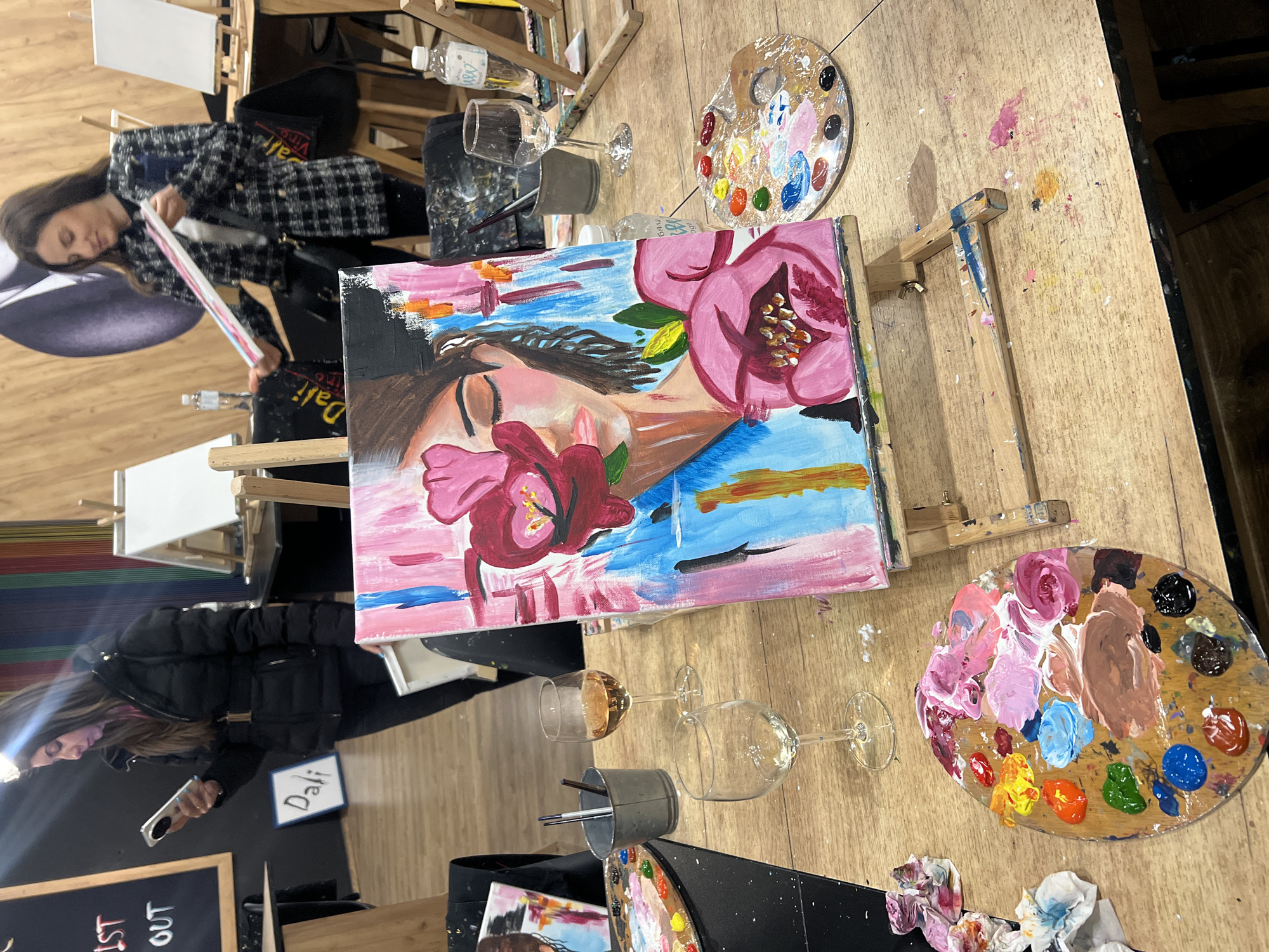The image depicts an indoor scene likely at an art class in Plovdiv, Bulgaria. Two women are present. One is painting, seen from behind, wearing a black and white jacket. The second woman is taking a photograph with her phone while wearing a black coat. In front of her is a sign with "Dali" written on it, along with a blackboard sign with some letters. In the foreground are several easels with partially completed paintings depicting a woman with flowers. There are also wooden palettes with various colors of paint, wine glasses, and containers with paintbrushes on the tables. The tables appear to be made of wood.
