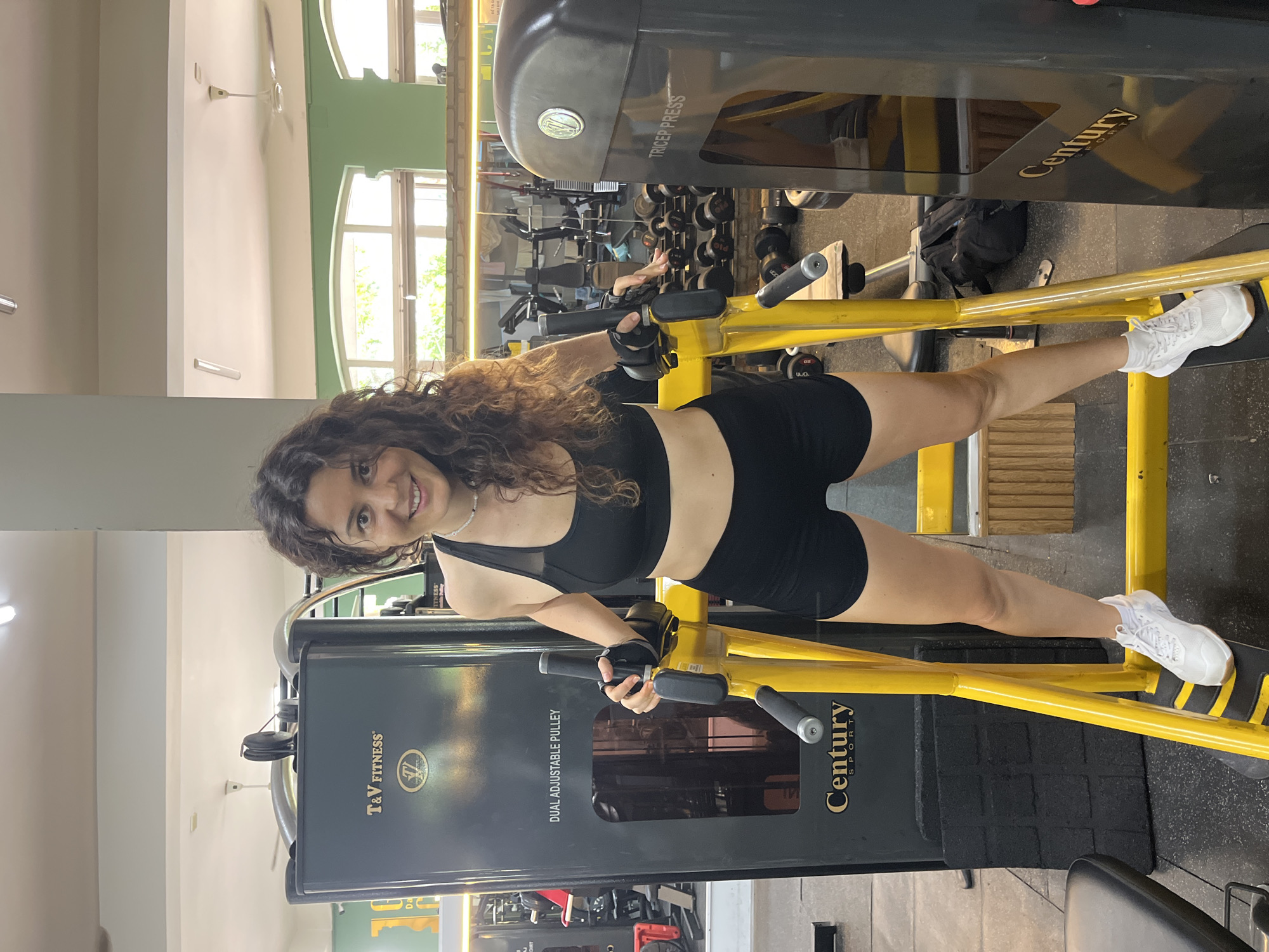 A young woman with curly brown hair, wearing a black sports bra and black shorts, stands in a gym in Phan Thiết, Vietnam. She is smiling and looking at the camera. She is positioned between two pieces of gym equipment. To her left is a black T&V Fitness Dual Adjustable Pulley machine, and to her right is a black Century Tricep Press machine. Both machines have yellow framing. She is holding onto the yellow handles of the Dual Adjustable Pulley machine. In the background, there are other gym machines, weights, and mirrors. The gym appears to be well-lit with natural light coming from windows.