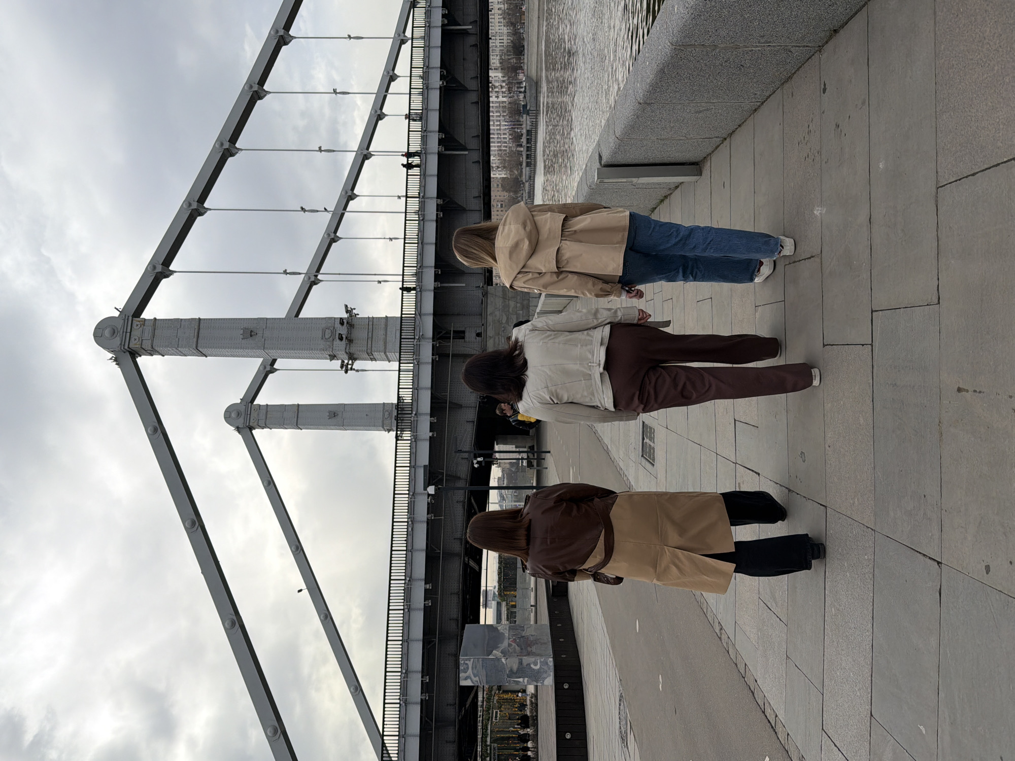 Three women are standing on a concrete walkway next to a river. They are facing away from the camera, toward the river and the bridge. The first woman is wearing a tan coat and blue jeans. The second woman is wearing a white jacket and brown pants. The third woman is wearing a brown coat and tan pants. The bridge appears to be the Crimean Bridge. The location is in the city of Moscow, Russia.