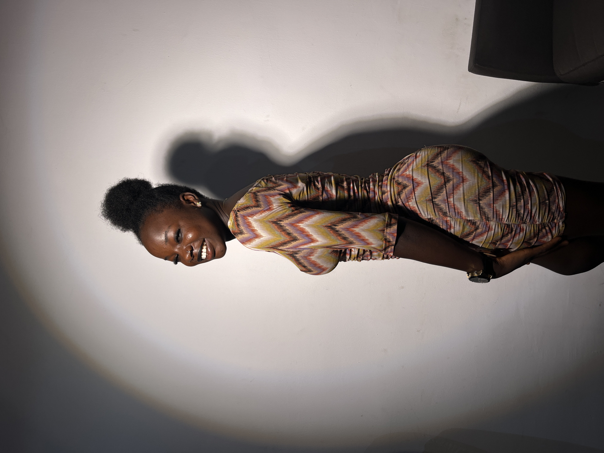 A young Black woman with dark skin and her hair in a bun is shown in a side profile, smiling at the camera. She is wearing a form-fitting, colorful, patterned dress with ruched detailing. She has on a watch on her left wrist and earrings. She is standing against a white wall with dramatic lighting creating a shadow of her body. A dark chair is partially visible on the right side of the frame.
