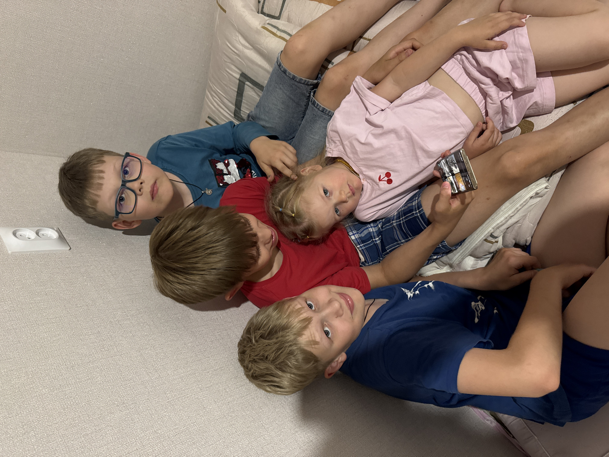 The photograph shows four children lying on a surface, possibly a couch or bed, with their heads angled towards the bottom left corner of the image. Starting from the top left, the first child wears blue glasses and a blue shirt with a graphic. The second child has brown hair and wears a red shirt. The third child, with blonde hair, is wearing a pink top and holding a small, metallic object (possibly a phone or small box). The fourth child has blonde hair and wears a blue shirt. They appear to be lying close together. The location may be Ферма, Russia. A white electrical outlet is visible on the wall. The background includes a beige wall and a patterned cushion.