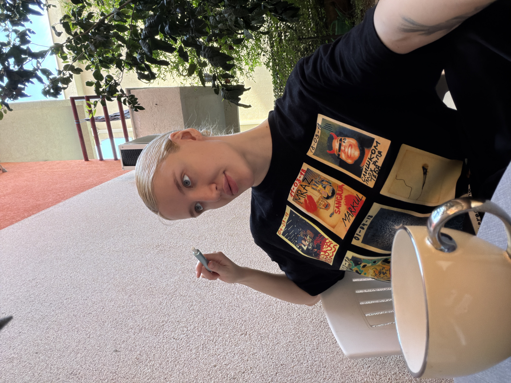 A fair-skinned woman with blonde hair and blue eyes is captured mid-expression, looking directly at the camera with a surprised or curious gaze. She is wearing a black t-shirt featuring several square graphics, resembling album covers or concert posters, with visible text including "16-03-1-", Cyrillic "СЛИШКОМ ПЛОХ ДЛЯ ТЕБЯ" (Too bad for you) alongside a person with devil horns, and "SANGAM MARKUL" with the date "01-04-18". Other dates like "04-03-18" and "11-04-18" are also visible on the shirt.
The woman holds a light blue/green object, possibly a vape pen or stylus, in her right hand. She is seated outdoors, with a white plastic chair partially visible next to her. In the immediate foreground, a cream-colored ceramic mug with a silver rim and handle is present.
The scene is an outdoor setting, likely a patio or balcony in Petrovac na Moru, Montenegro, under bright daylight. Lush dark green foliage from a tree or shrub hangs overhead, casting shadows. In the background, light-colored buildings and distinct red/maroon vertical structures can be seen. The ground surface features a light-colored, speckled texture in the foreground, transitioning to a reddish-brown area on the left. The photo is angled, suggesting a casual, spontaneous shot.