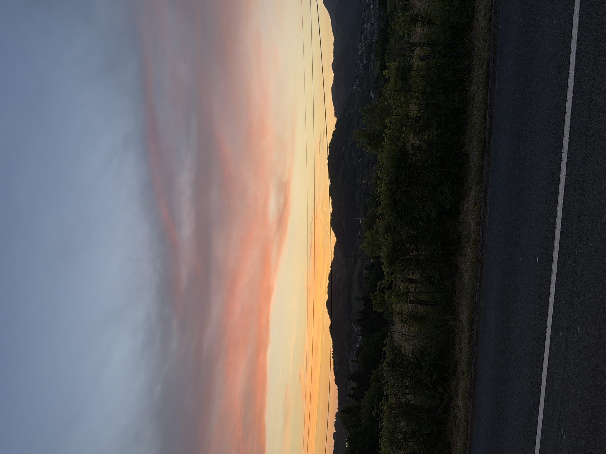 The image depicts a landscape at dusk or dawn, likely in a rural or semi-rural area. A road with a white line marking runs along the right side of the frame. Adjacent to the road is a strip of vegetation. Beyond the vegetation is a dark, hilly landscape. The sky is a mix of colors, including yellow, orange, and pink, suggesting a sunset or sunrise. Power lines are visible. It's possible to locate the exact spot along the GI-3681 if additional landmarks are visible, specifically if the position of the town on the top of the mountain can be linked to a road location.