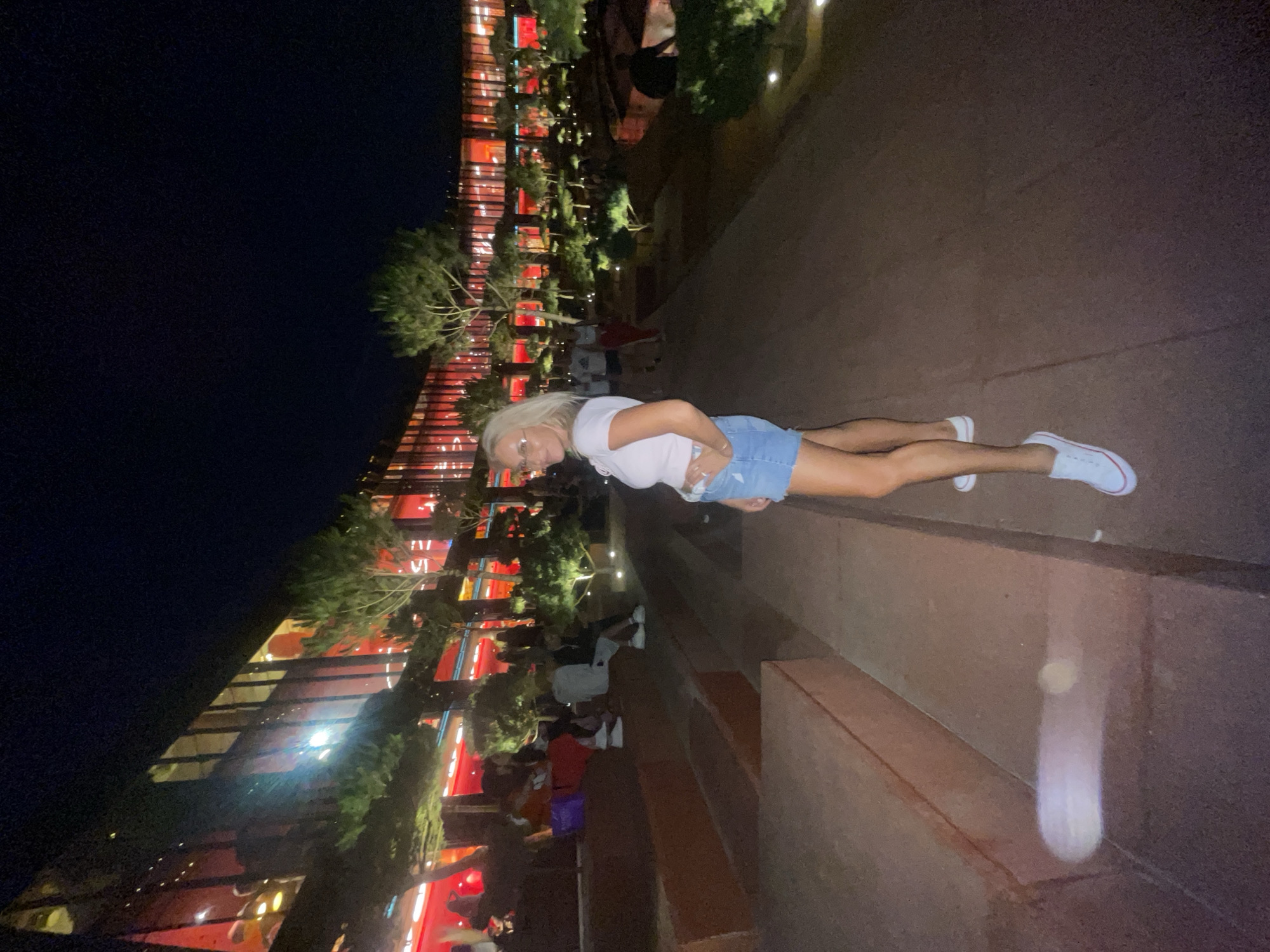 In this vertically oriented nighttime image, a group of people are gathered outdoors in what appears to be a public space. A woman with blonde hair and glasses, wearing a white t-shirt and a denim skirt, is the primary subject in the foreground. She is lying on her side, facing the camera. Behind her, a modern building with red illuminated windows and balconies is visible, surrounded by trees. The building's architecture suggests a contemporary design, possibly a hotel or a cultural center. Several other individuals are scattered throughout the background, some seated and others standing, creating a sense of a social gathering or event. The lighting suggests it is evening or night, with artificial lights from the building and scattered lights in the trees illuminating the scene. The ground in the foreground appears to be a textured, dark surface, possibly stone or concrete, with some subtle lighting effects. The overall atmosphere seems relaxed, with people enjoying the nighttime ambiance. The location is identified as Krasnodar, Russia. No visible text is present in the image.