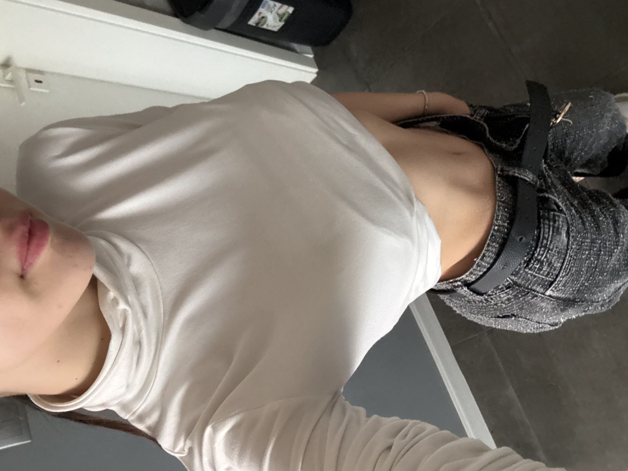 The image shows a person wearing a white turtleneck sweater and dark denim shorts with a black belt. The person is positioned near a white surface that appears to be a counter or shelf, and the background suggests an indoor setting, possibly a bathroom or utility area. The person's face is partially visible, showing the lips and a portion of the cheek. Without additional context or landmarks, it is impossible to determine the specific location in Sofia, Bulgaria.