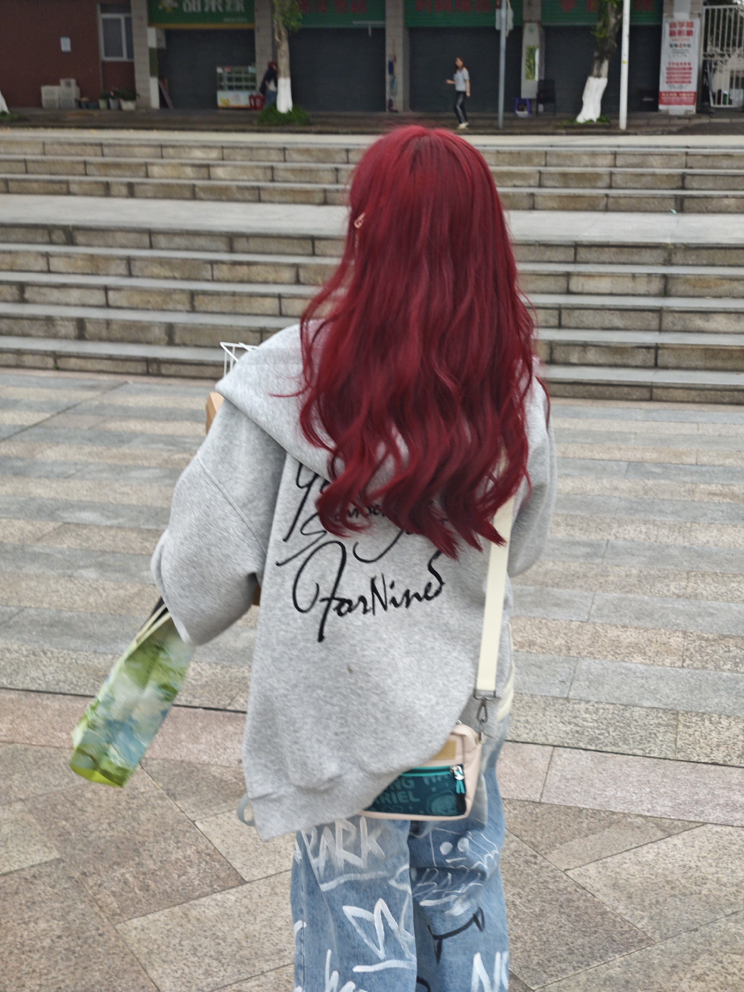The image shows a person with long, wavy red hair, wearing a gray hoodie with black lettering, blue jeans with white doodles, and a small beige purse with a teal detail on the front. The person is holding a plastic bag with a greenish-blue design. The person is seen from the back, standing on a paved area with rectangular tiles. In the background are stairs leading up to a street level with shops and trees. People are walking on the street. Based on the architecture, signage, and overall environment, the location is likely in an urban area in the city of Chongqing High-tech Zone, China.