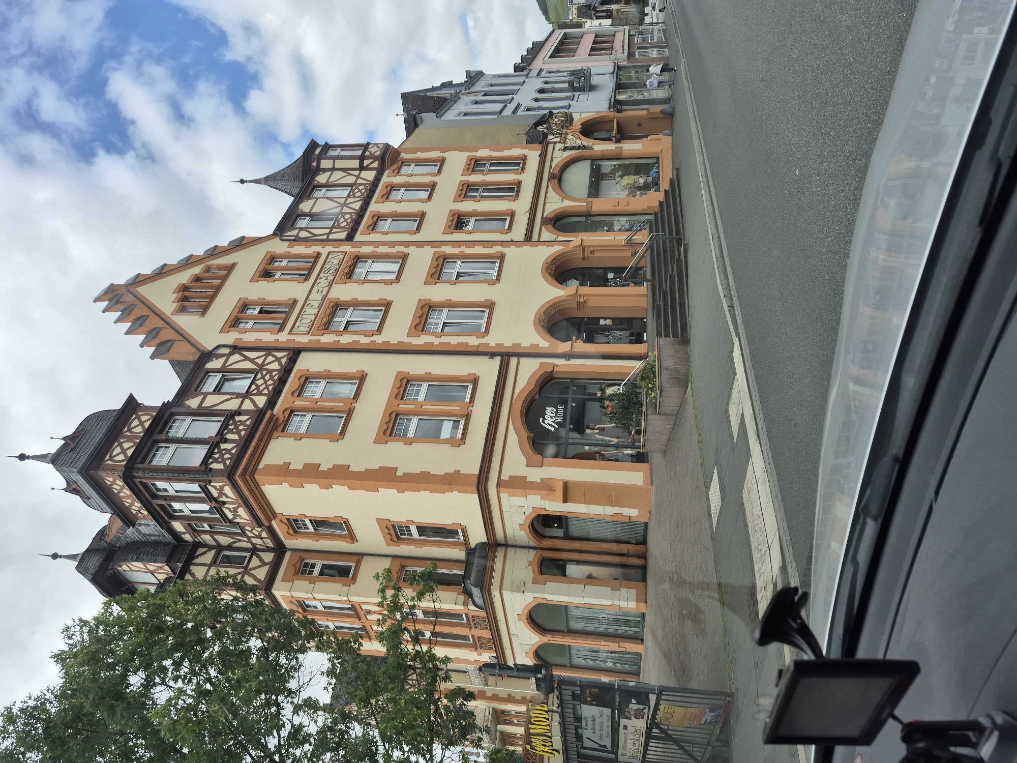 This image depicts a street scene in Bernkastel-Kues, Germany, specifically facing the Hotel Gassen at Cusanusstraße 7. The perspective is from inside a vehicle, looking forward and slightly to the right.
The most prominent structure is a large, multi-story building on the left, identified as "HOTEL-GASSEN" by lettering on its facade. This building features a light yellow-cream primary color with pronounced orange-brown architectural trim around windows, corners, and as decorative elements. The architecture includes traditional half-timbered sections on upper floors and gables, with dark grey slate roofs topped by multiple ornate spires and turrets. The ground floor of this building contains several arched storefronts and entrances; one prominent shop, "hees MODE," is visible on the far left with display windows showing mannequins. Steps lead up to several of the entrances from the sidewalk.
To the right of the Hotel Gassen, a row of interconnected buildings extends down the street. These structures exhibit varying pastel colors, including light blue, pink, and grey, and also feature traditional European architectural styles, some with half-timbering and decorative elements. Further down the street, two indistinct individuals are walking on the sidewalk away from the viewer.
The street in the foreground is asphalt, with a clear curb separating it from the sidewalk. White rectangular markings of a crosswalk are visible on the pavement. Reflections are present on the dashboard and lower windshield of the vehicle from which the photo was taken. A black navigation device is mounted on the dashboard in the lower-left corner. On the far left, partially obscured by a tree, a metal fence and a signpost are visible, with one sign mentioning "Brückenkeller."
The sky is partly cloudy, with significant grey cloud cover but also clear patches of blue sky visible, particularly towards the upper right. A tree with green foliage partially obscures the upper left portion of the main hotel building. In the distant background to the right, a vegetated hillside is visible, indicative of a river valley landscape.