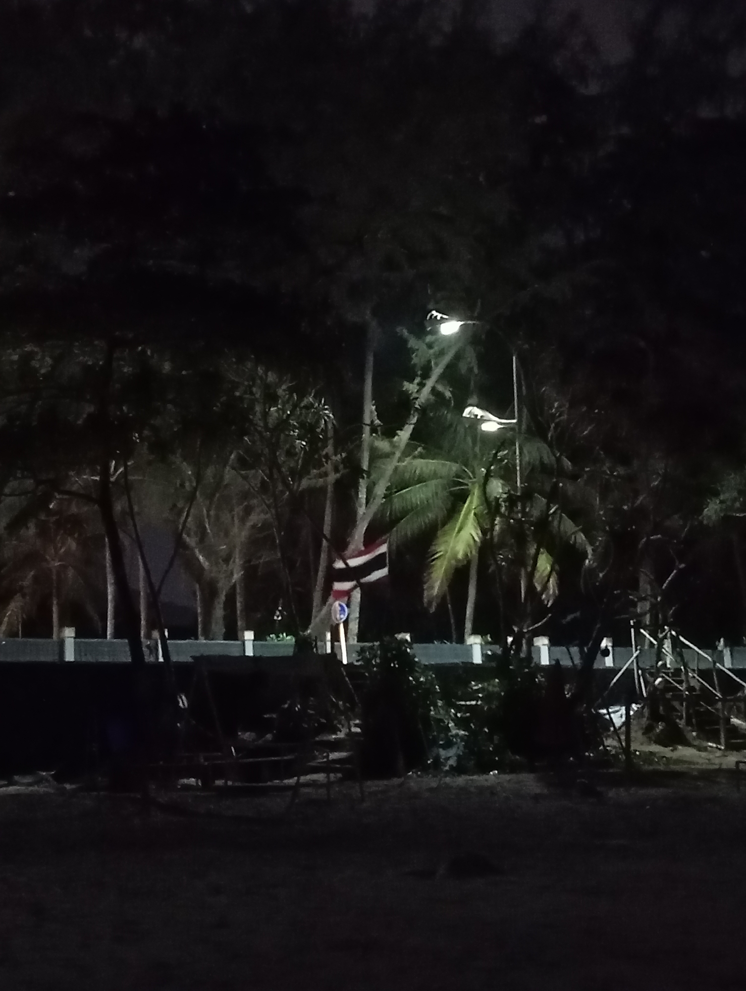 A nighttime photograph captures a dimly lit beach scene in Karon, Thailand. In the background, a row of white bollards marks a boundary, with palm trees and other vegetation beyond. Two streetlights illuminate the area, casting a soft glow. A Thai flag is visible, attached to a pole. The foreground shows a sandy area with some scattered debris and structures that appear to be made of wood and metal, possibly remnants of playground equipment or shelters. The overall atmosphere is dark and somewhat desolate, suggesting a quiet, off-season or late-night setting.