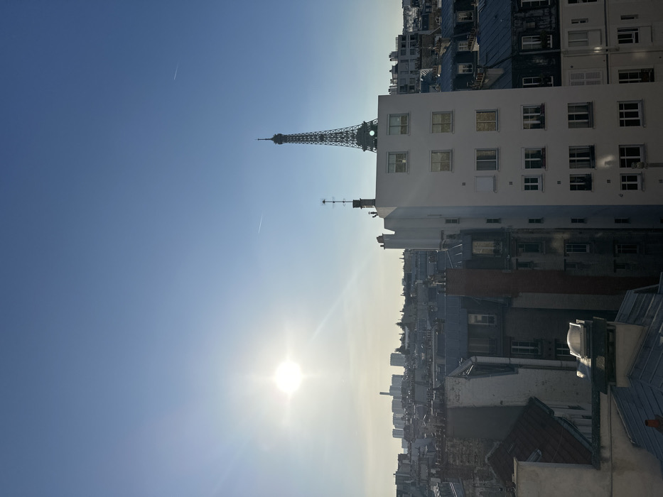 The image displays an urban panorama of Paris, France, captured from an elevated perspective during daytime. A clear, bright blue sky occupies the upper portion, featuring a prominent solar disc with strong lens flare in the upper-left quadrant and two faint aircraft contrails. The Eiffel Tower is visible in the upper-right quadrant, its upper structure rising above a prominent, light-colored, multi-story building in the midground. This building exhibits multiple rectangular windows and is topped with a chimney featuring an antenna. The foreground and midground are composed of a dense arrangement of traditional Parisian buildings, characterized by their grey, pitched zinc roofs and numerous chimneys. A distant cluster of high-rise buildings forms a hazy skyline against the horizon in the left background. No individuals or specific activities are discernible within the frame.