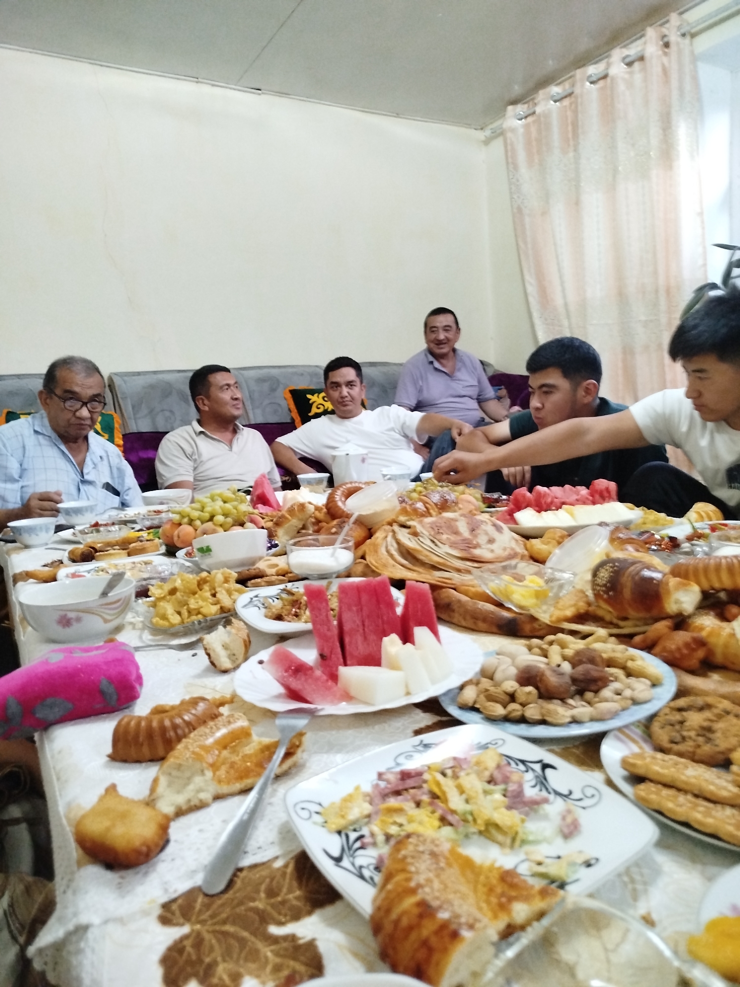 A group of five men are seated around a table laden with food, likely for a meal or celebration. The setting appears to be indoors, with a pale wall and patterned curtains visible in the background. The men are of varying ages, with some appearing to be elderly and others younger. They are dressed casually. The table is generously covered with a variety of food items, including pastries, bread, fruits (grapes and what appears to be watermelon), nuts, and some dishes that are not clearly identifiable but seem to include salads and possibly fried items. The atmosphere seems relaxed and communal. The specific location is the city of Кызыл-Булак, Kyrgyzstan.
