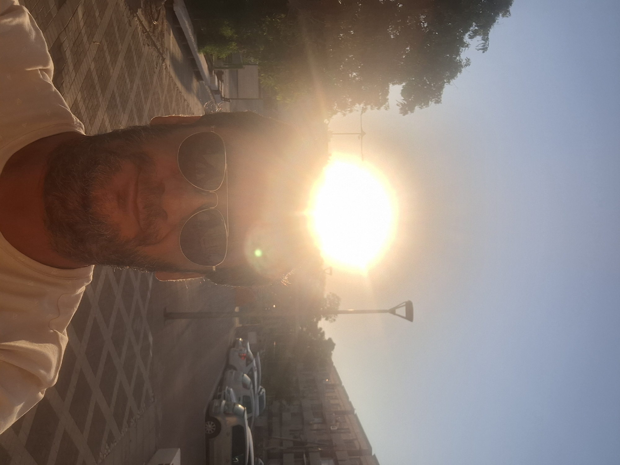 A close-up, eye-level, selfie-style photograph captures a man in his mid-40s, with dark hair and a beard, wearing aviator sunglasses. He is positioned in the foreground, with the bright, setting sun directly behind his head, creating a halo effect. The man is outdoors, appearing to be on a paved walkway in an urban environment. In the background, to the left, are trees with green foliage. To the right, a street is lined with parked cars, and beyond that, a multi-story building with numerous windows. A streetlamp is visible in the mid-ground, to the right of the sun. The overall lighting suggests it is either sunrise or sunset. The man is wearing a light-colored t-shirt. The scene is likely the city of Yerocham, Israel, given the typical urban landscape and vegetation.