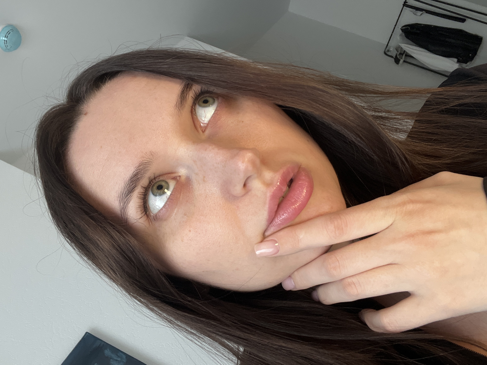 A close-up, angled shot features a young woman with light skin, brown hair, and green eyes. She is looking upwards with her lips slightly parted and glossy. Her fingers are lightly touching her chin. In the background, portions of a white wall, a smoke detector, and a clothes rack with hanging items are visible. The location is not determinable as being in Tyumen, Russia based on the image.