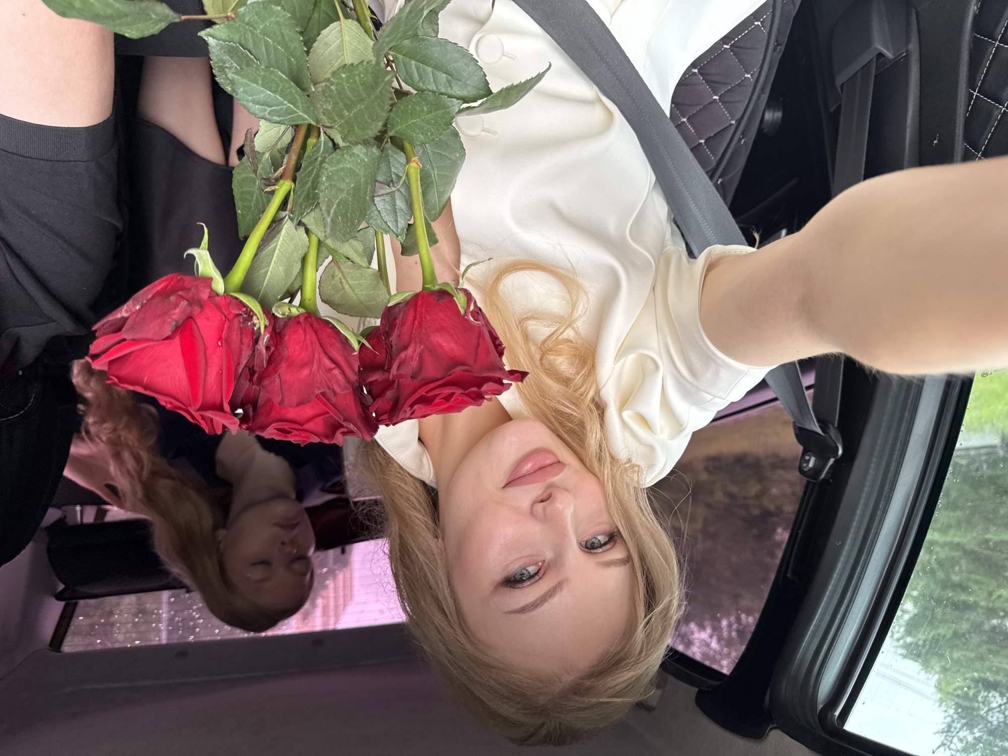 A young woman with blonde hair and blue eyes is taking a selfie in the passenger seat of a car. She is holding a bouquet of red roses. She is wearing a white blouse. Behind her, another young woman with blonde hair is visible in the back seat, appearing to be asleep. The car appears to be parked or moving slowly, as indicated by the view outside the window, which shows trees and foliage. The interior of the car is visible, including a seatbelt and quilted seat covers. The overall lighting suggests it is daytime, possibly overcast. The location is the city of Perm, Russia.