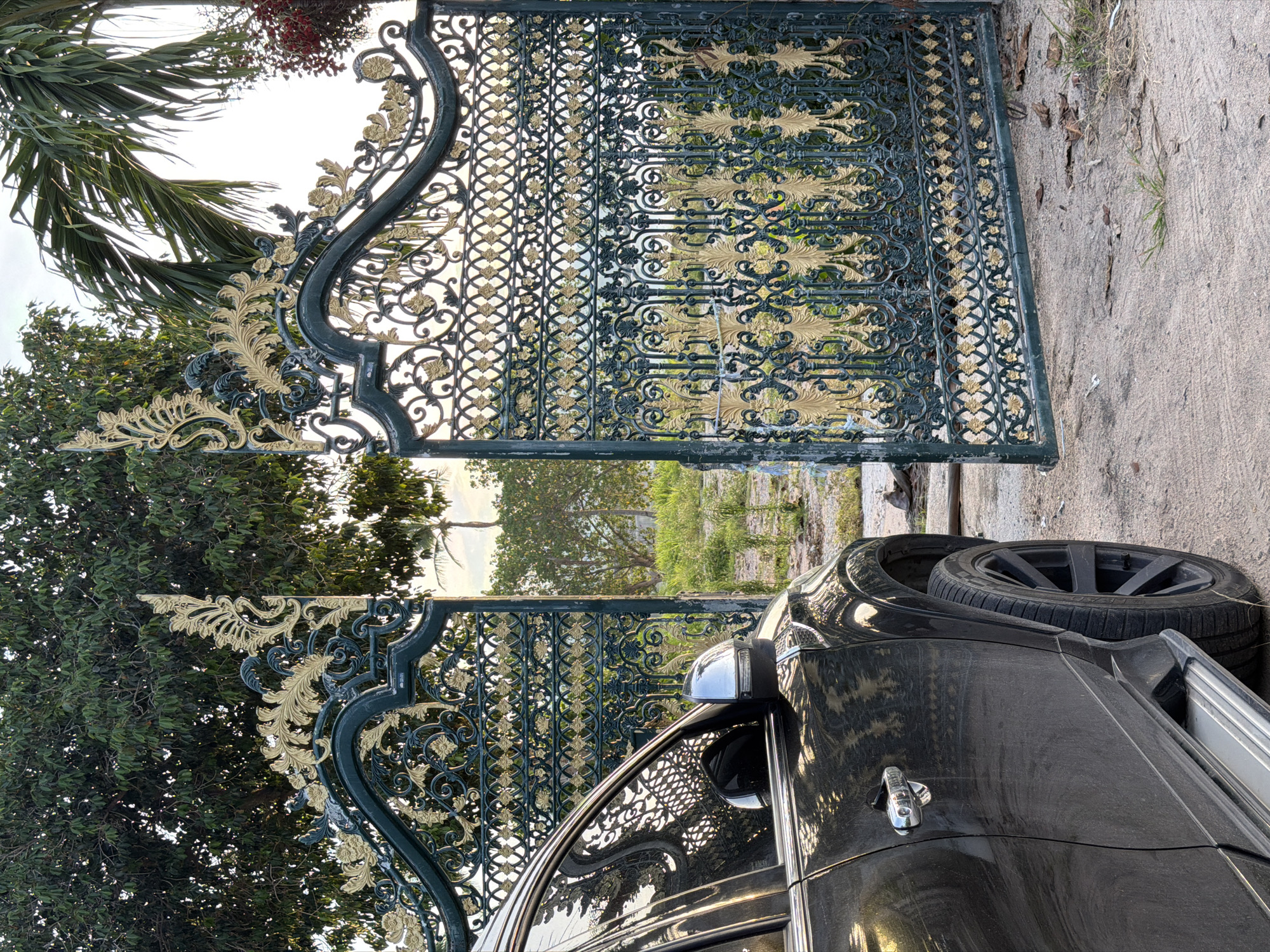 The image depicts a black SUV parked next to an ornate, dark green metal gate with gold detailing. The gate is a focal point, featuring intricate scrollwork and floral motifs, and is partially open, revealing a glimpse of greenery and trees beyond. One section of the gate is open, allowing the car to be positioned beside it. The car itself is a dark color, reflecting the surroundings and the sky. The wheel of the car is visible, showing a modern alloy rim.
The setting appears to be outdoors, with a sandy or gravelly ground surface next to a concrete path. Lush green trees and foliage are visible behind the gate, suggesting a tropical or subtropical environment. A palm frond can be seen in the upper left corner of the image. The lighting suggests it could be daytime, possibly late afternoon, with a bright sky. There are no people visible in the image, nor any text. The scene evokes a sense of entering or exiting a property, with the grand gate suggesting a place of significance, perhaps a private residence or a special establishment in Baan Plai Laem, Thailand.