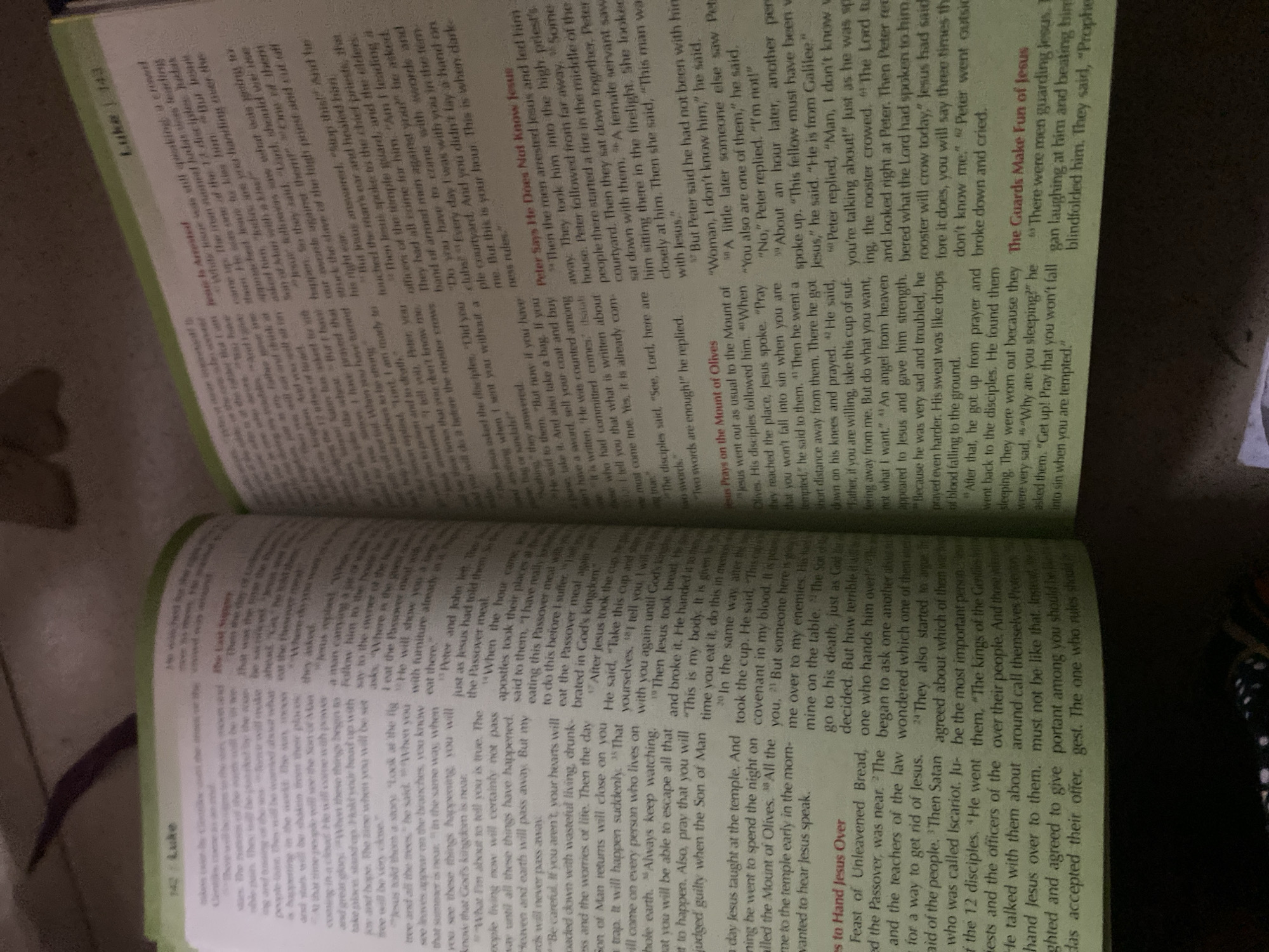 The image shows an open Bible, with text visible on both pages. The left page displays the heading "es to Hand Jesus Over" and a section of text in a smaller font, accompanied by numbered verses. The right page is filled with text in a smaller font, also with numbered verses, and includes a heading in red that reads "The Guards Make Fun of Jesus." The lighting in the image suggests it was taken indoors, possibly in a study or library, with the Bible resting on a surface. The perspective is from slightly above and to the side, capturing the detail of the printed text and the book's binding. There is no discernible connection to Jauro Yinu, Nigeria, or any elements that would indicate a specific urban location.