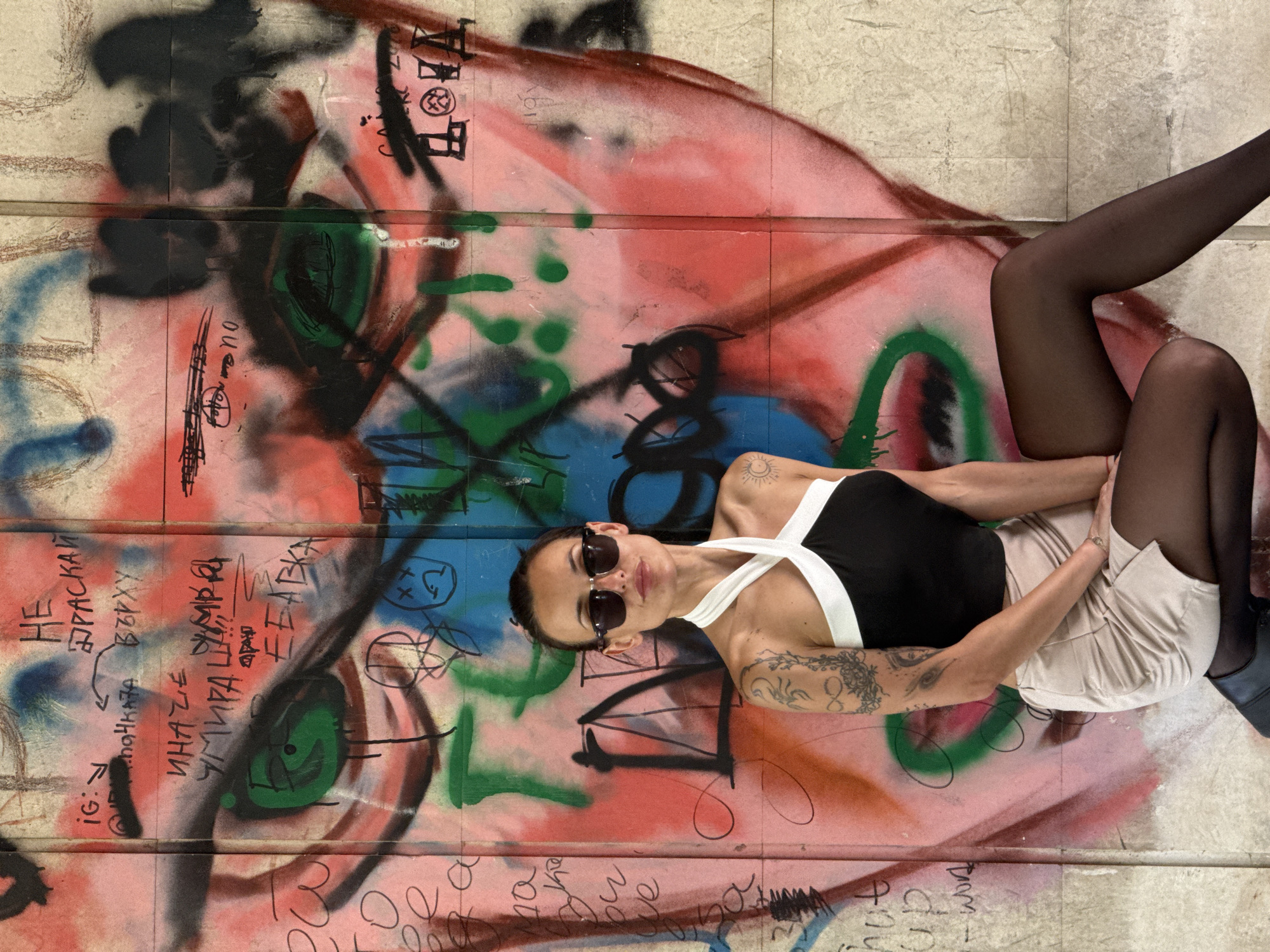 The image shows a woman lying on a painted wall. She is wearing a black and white top, a beige skirt, black stockings, and sunglasses. Her arms are tattooed. The wall features graffiti in various colors, including red, green, blue, and black. Some graffiti appears to be in Bulgarian. A city of Pleven in Bulgaria is mentioned.