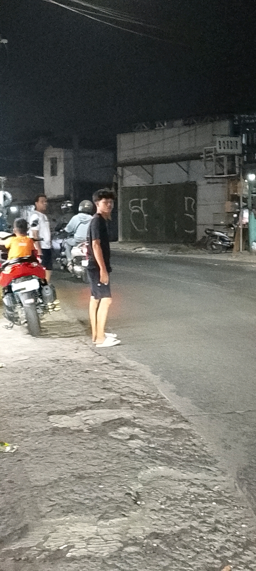 In the city of Karangsatria, Indonesia, at night, a young man with curly hair stands on the side of a road. He is wearing a black t-shirt and dark shorts, with white flip-flops on his feet. To his left, a red motorcycle is parked. Further down the road, another motorcycle is visible with a person on it wearing a light blue jacket and a helmet. To the far left, a person in an orange shirt is seated on a motorcycle. Another man in a white shirt stands near the orange-shirted individual. The background features a building with a light-colored facade and a dark wall with graffiti. The road surface is paved and appears wet in some areas.