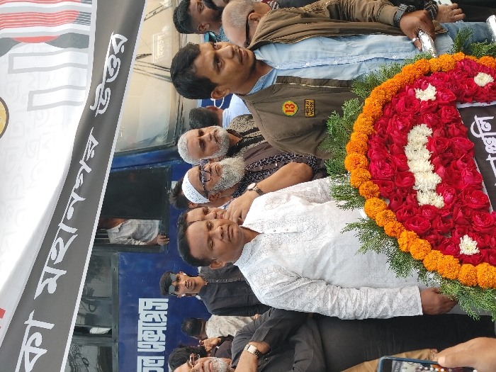The image displays a daytime outdoor gathering of approximately a dozen men in Dhaka, Bangladesh, specifically within the Dhaka Metropolitan North area, as indicated by a prominent banner. In the immediate foreground, a man in a white, intricately embroidered long-sleeved kurta stands centrally, facing forward with a neutral expression. To his right, a younger man in a brown bomber jacket over a light blue shirt holds a large, circular floral wreath with both hands. The wreath is composed primarily of red roses, accented by a band of white flowers, bordered by orange marigolds, and interspersed with green foliage. A black banner with white Bengali script is visible beneath the wreath, while another, larger black and white banner bearing the Bengali text "ঢাকা মহানগর উত্তর" (Dhaka Metropolitan North) stretches horizontally across the upper portion of the frame, behind the gathered individuals.
Behind the central figures, a crowd of additional men is visible. These include an individual with a white beard and white kufi (prayer cap) wearing glasses, another man with a grey beard, and a younger man wearing a black shirt and sunglasses. In the background, a blue bus is partially visible, with a person looking out from one of its windows. Bengali script, possibly "অকাল এক্সপ্রেস," is discernible on the side of the bus. On the far left, a hand holding a mobile phone is actively taking a photograph of the scene, partially obscuring the left side of the man in the white kurta. The overall setting is an urban outdoor environment.