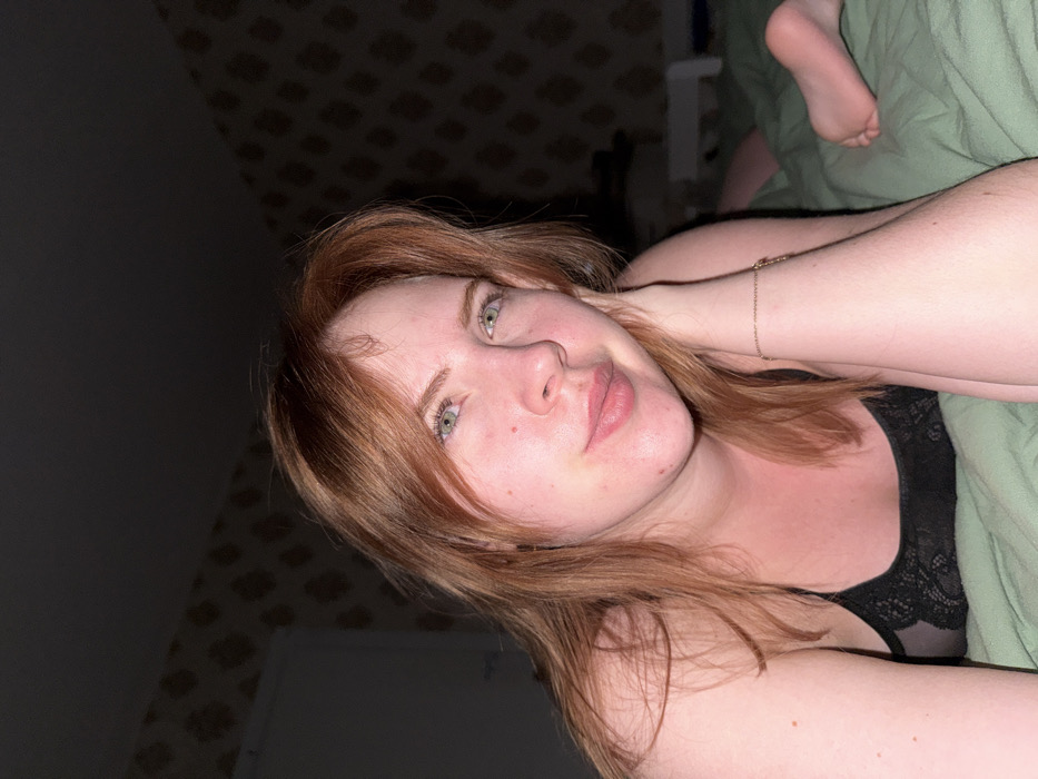 A woman with fair skin and red hair lies on a green bed. She's wearing a black lace top, with a delicate gold bracelet on her raised arm. Her eyes are green. Her expression is relaxed. The background is partially a dark wall with a patterned covering. A foot is visible near the top of the frame.