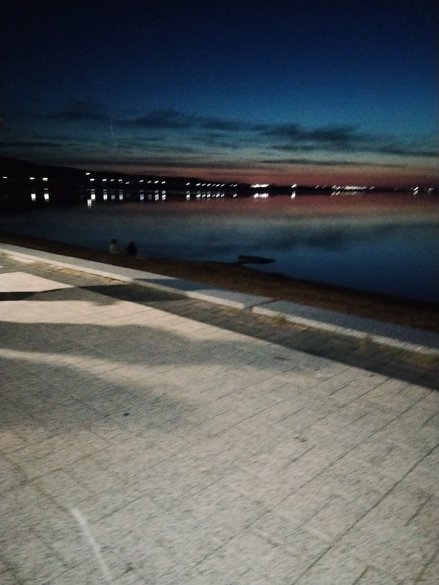 The image depicts a waterfront scene at dusk or night in Kokshetau, Kazakhstan. A paved walkway with patterned blocks occupies the foreground, casting long shadows. Beyond the walkway, a body of water reflects distant lights, suggesting an urban or semi-urban setting along the shore. The sky transitions from deep blue at the top to pinkish-orange hues near the horizon, indicating the last light of day or early twilight. On the far shore, a line of illuminated structures is visible, their lights creating reflections on the water. In the middle ground, near the water's edge, two indistinct figures are seated, facing the water. The overall impression is one of a tranquil evening by the water.
