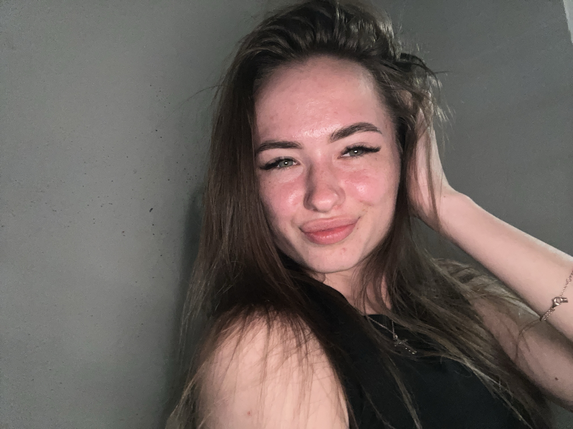 A young adult female poses for a close-up indoor portrait against a plain, dark gray wall in the city of Unknown, Unknown. She has long, straight brown hair, some strands falling across her forehead and framing her face. Her right hand (viewer's left) is raised, with fingers lightly touching the side of her head and running through her hair. Her complexion appears flushed or reddish on her forehead and cheeks, showing visible skin texture and small blemishes. She has full, dark eyebrows and green or hazel eyes enhanced with prominent dark eyelashes. Her lips are slightly pursed in a 'pouty' expression, with a subtle upward curve at the corners. She wears a black sleeveless top, revealing her left shoulder (viewer's right), and accessorizes with a delicate silver-toned necklace featuring a small cross-shaped pendant, and a thin, possibly rose gold, chain bracelet with a small key or lock charm on her right wrist (viewer's left). The lighting is soft, illuminating her face and creating minor shadows, suggesting an artificial light source.