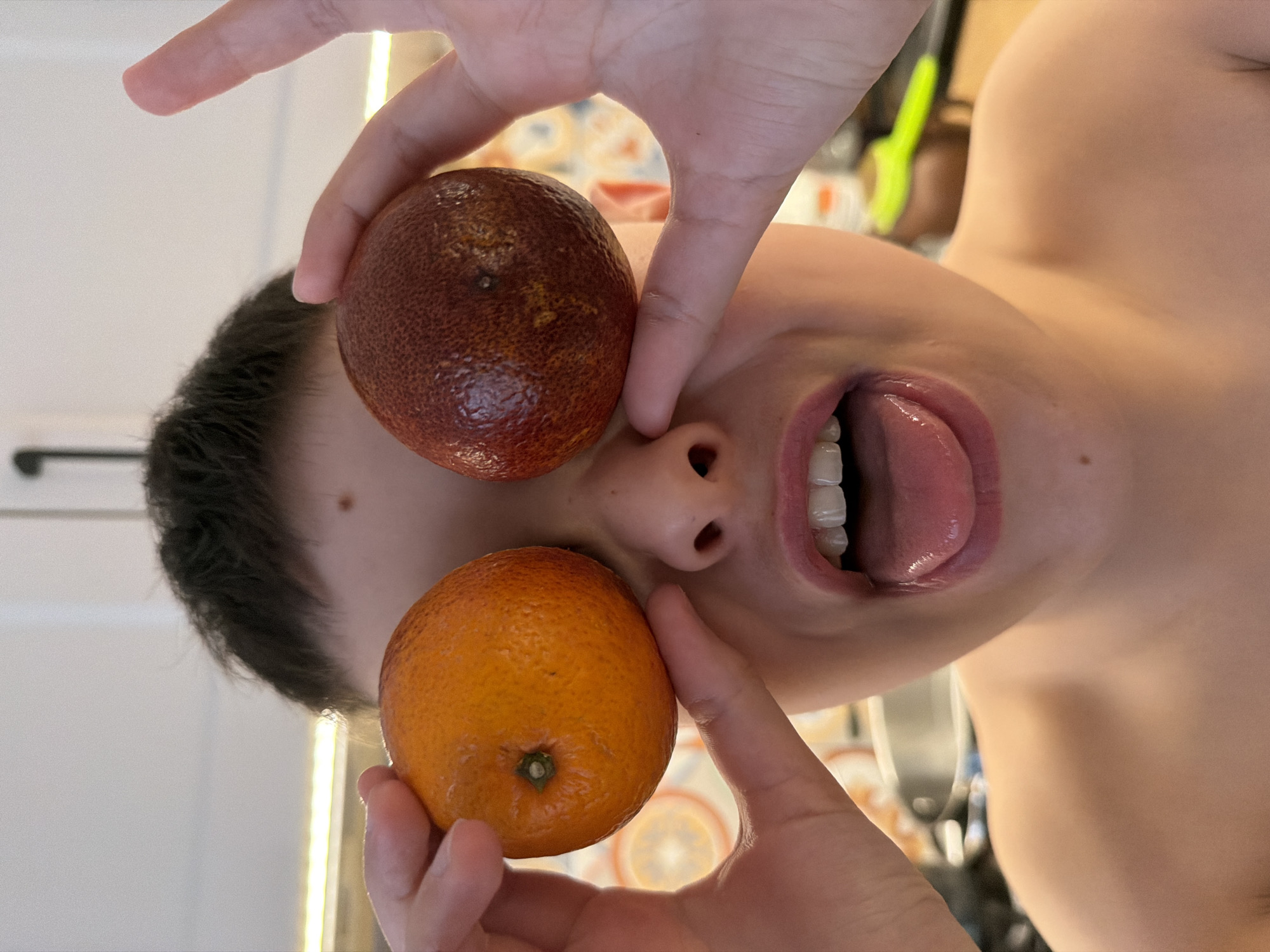 A person lies on their back, holding a blood orange over one eye and a tangerine over the other, obscuring them from view. The person's mouth is open with their tongue extended. The background shows a partial view of what appears to be a kitchen setting. The location of this image is not deducible from the photograph.