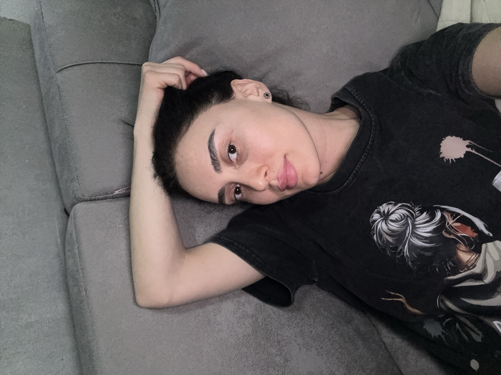A woman lies on a gray couch. She is fair-skinned and has dark hair. She is wearing a dark t-shirt. Her right arm is bent, and her hand is near her head. The location is not discernible from the image.