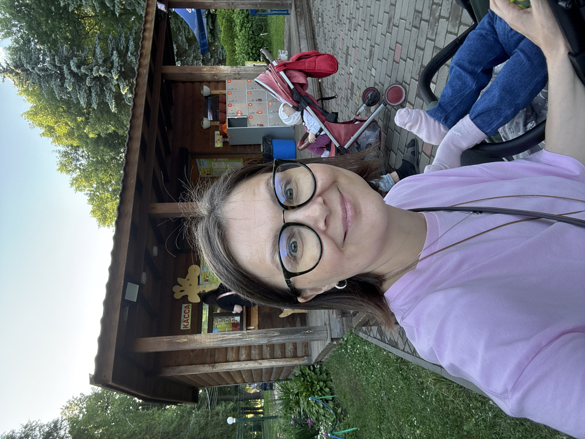 A woman with brown hair and glasses takes a selfie in front of a wooden kiosk with "KACCA" signage and a giraffe graphic. The kiosk appears to be a ticket booth or shop. Behind her, a baby is seated in a red stroller. The scene is outdoors, with trees and grass visible, suggesting a park or recreational area. The paving stones indicate a walkway. The presence of lockers and a blue trash can suggests amenities for visitors. The overall setting is a daytime scene in a public outdoor space.