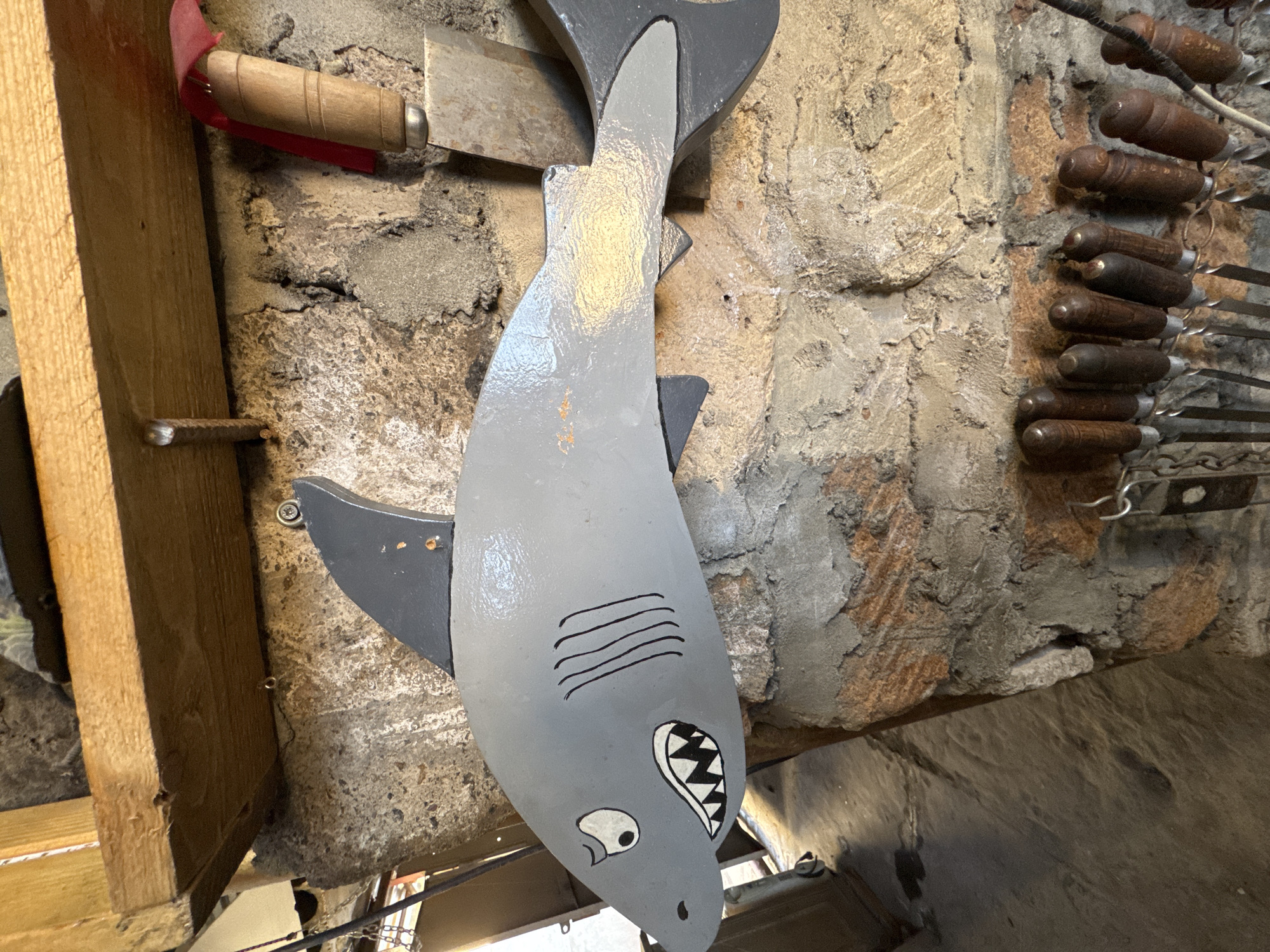 The image captures a rustic indoor or covered outdoor setting, possibly a kitchen or barbecue area, dominated by a decorative, painted wooden shark hanging vertically on a textured wall.
The primary subject is a stylized, light gray wooden shark figure. It features darker gray fins and tail, black painted gills, a white eye with a black pupil, and an open mouth revealing jagged white and black teeth. Faint golden reflections are visible on its body, suggesting a glossy finish or specific lighting.
The background consists of a rough, uneven wall, appearing to be constructed from stone or concrete with patches of plaster in various shades of gray, beige, and reddish-brown. Some areas show exposed, weathered rock textures.
To the left and slightly behind the shark's head, a meat cleaver or chopper is visible, featuring a light wooden handle wrapped with a bright red fabric or ribbon, and a dull metallic blade. On the right side of the wall, a rack holds approximately nine metal skewers, each with a dark, textured wooden handle. A vertical wooden beam or plank runs along the far left edge of the frame, and a small metal pin or nail protrudes from the wall near it.
No activity is taking place; the objects are static and serve as decor or stored tools. The scene evokes a rustic and functional ambiance. There is no visible text in the image.