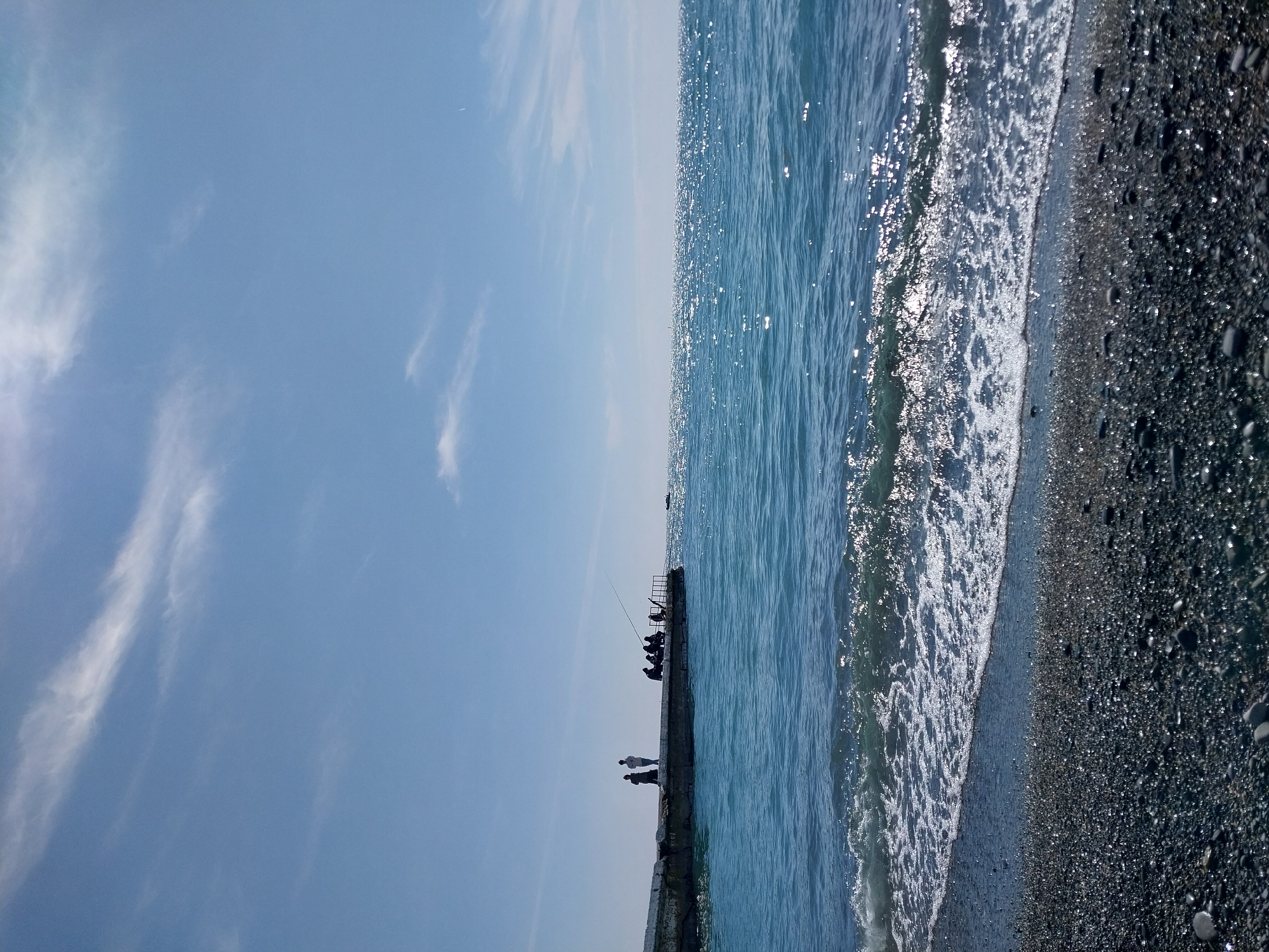 This image captures a vibrant coastal scene in Sochi, Russia, on a bright, sunny day. The composition is divided, featuring a clear sky on the left, and the sea, a pier, and a pebbly beach on the right (if viewed in its intended landscape orientation after rotation).
The sky, occupying roughly half of the frame, is a brilliant light blue with soft, wispy white clouds stretching across it, indicating fine weather. The strong sunlight illuminates the entire scene.
On the right, the sparkling blue sea dominates, its surface intensely shimmering with sunlight, creating numerous bright reflections. Gentle waves lap against a dark, pebbly beach, producing lines of white foam along the waterline. The beach consists of small, dark, glistening stones.
Extending into the water from the left side is a dark, low-lying pier or breakwater. Several people are silhouetted against the bright backdrop of the sea and sky. In the foreground section of the pier, two distinct figures can be seen standing. Further along the pier, a group of at least five individuals are gathered, and at least one fishing rod is visible, suggesting they are fishing. Their features are indistinguishable due to being silhouettes.
The overall impression is one of a calm, recreational day at the seaside. There is no visible text in the image.