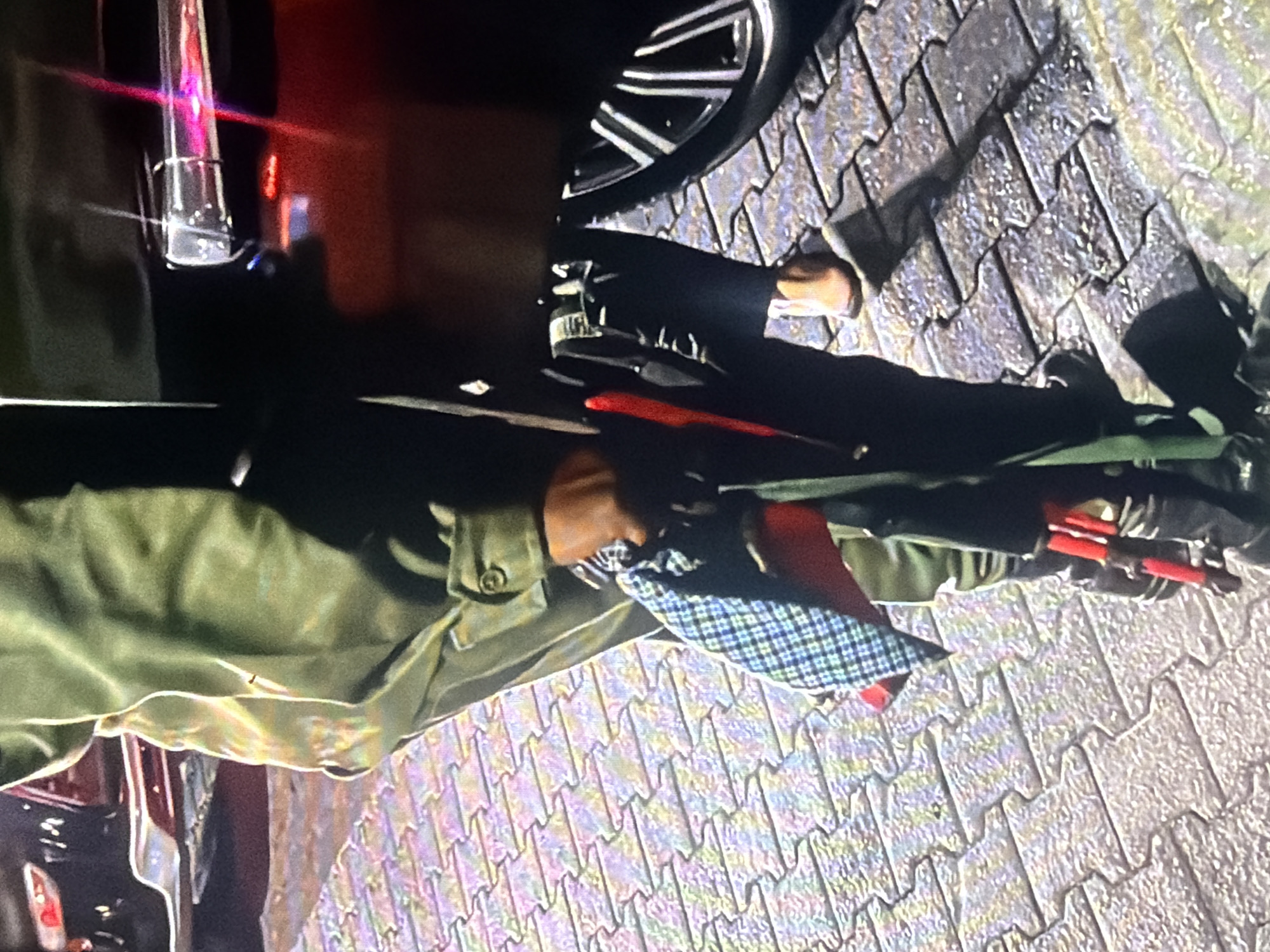 The image shows a close-up view of a person holding several items, including a checkered umbrella, a red object, and a black bag. The person is wearing a green garment and dark pants. They are standing on a patterned paved surface, possibly a sidewalk or courtyard. In the background, parts of a dark-colored vehicle are visible, including a wheel and a door handle. The lighting suggests it might be evening or a dimly lit indoor space. The specific location in Bukuru, Nigeria, is not discernible from the visual information provided.