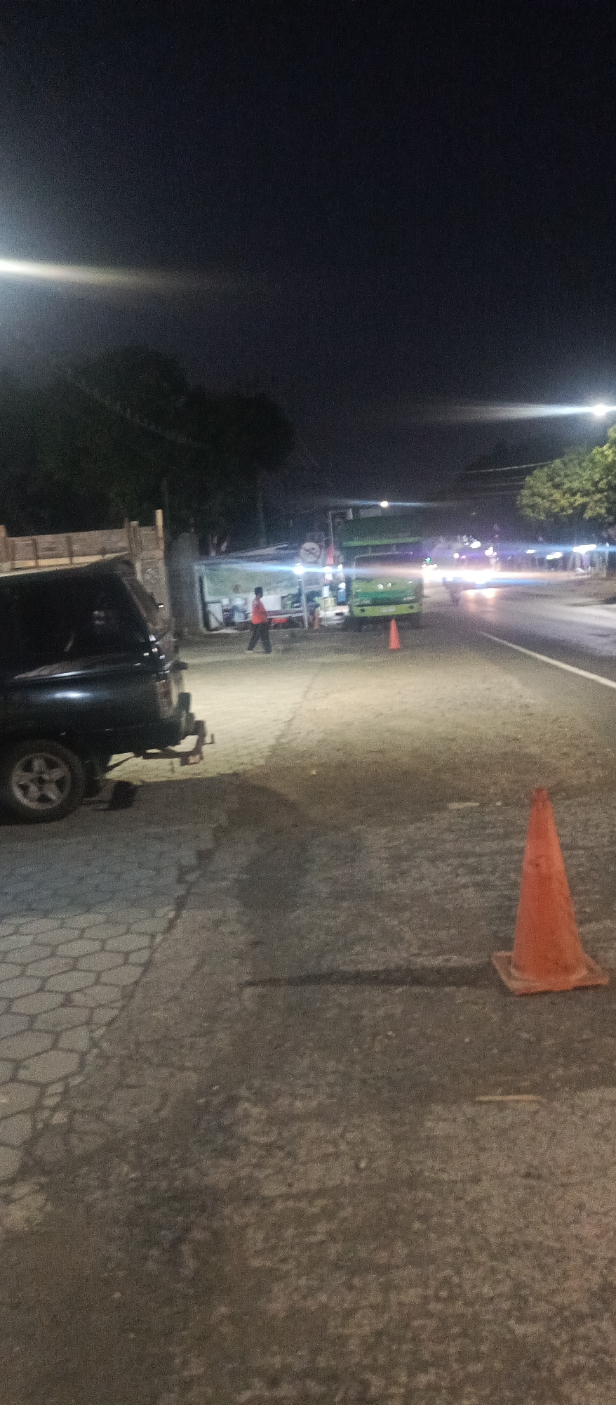 This is a night-time vertical photograph taken from street level in Arjasa, Indonesia. The scene depicts a street with several vehicles and a person. In the foreground on the left is the rear of a dark-colored SUV, with its rear wheel clearly visible. The ground in the foreground is paved with interlocking hexagonal stones, transitioning to asphalt pavement further into the scene. An orange traffic cone is placed on the asphalt on the right side of the image. In the middle ground, a person wearing an orange shirt is walking. Behind the person are several stalls or small shops, one of which has a green and white awning. A large green truck is parked or stopped in front of these stalls. Further down the street, headlights from oncoming traffic are visible, suggesting the street is open for vehicles. The sky is dark, indicating it is nighttime, with a bright light source, likely a streetlamp, visible in the upper left quadrant of the image. Trees and vegetation are present in the background, silhouetted against the night sky.